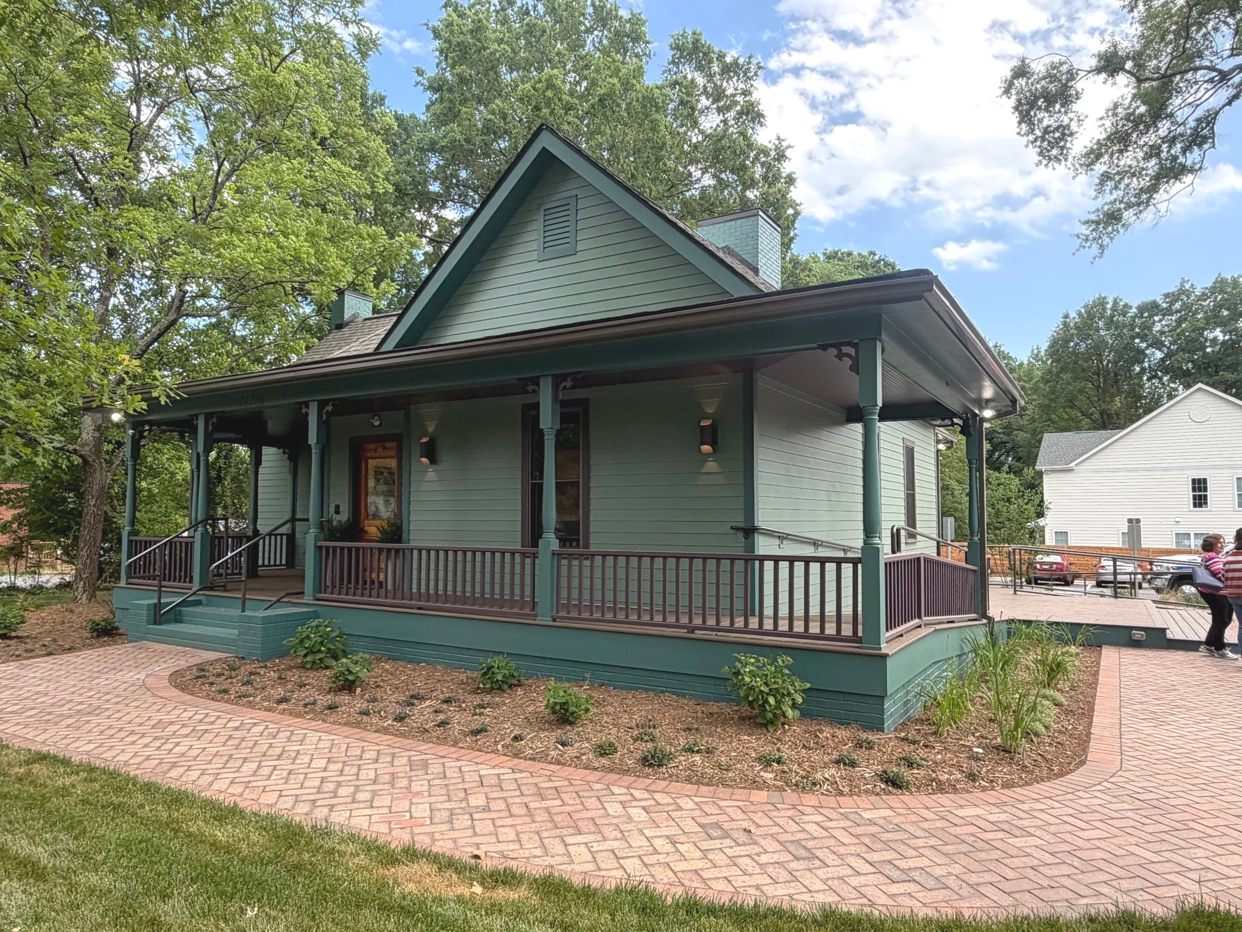 A two-story house painted with light teal color featuring a front porch with a railing, dark railing and stairs, and a gabled roof. The house is surrounded by green trees and bushes. The sky is partly cloudy with patches of blue showing through.