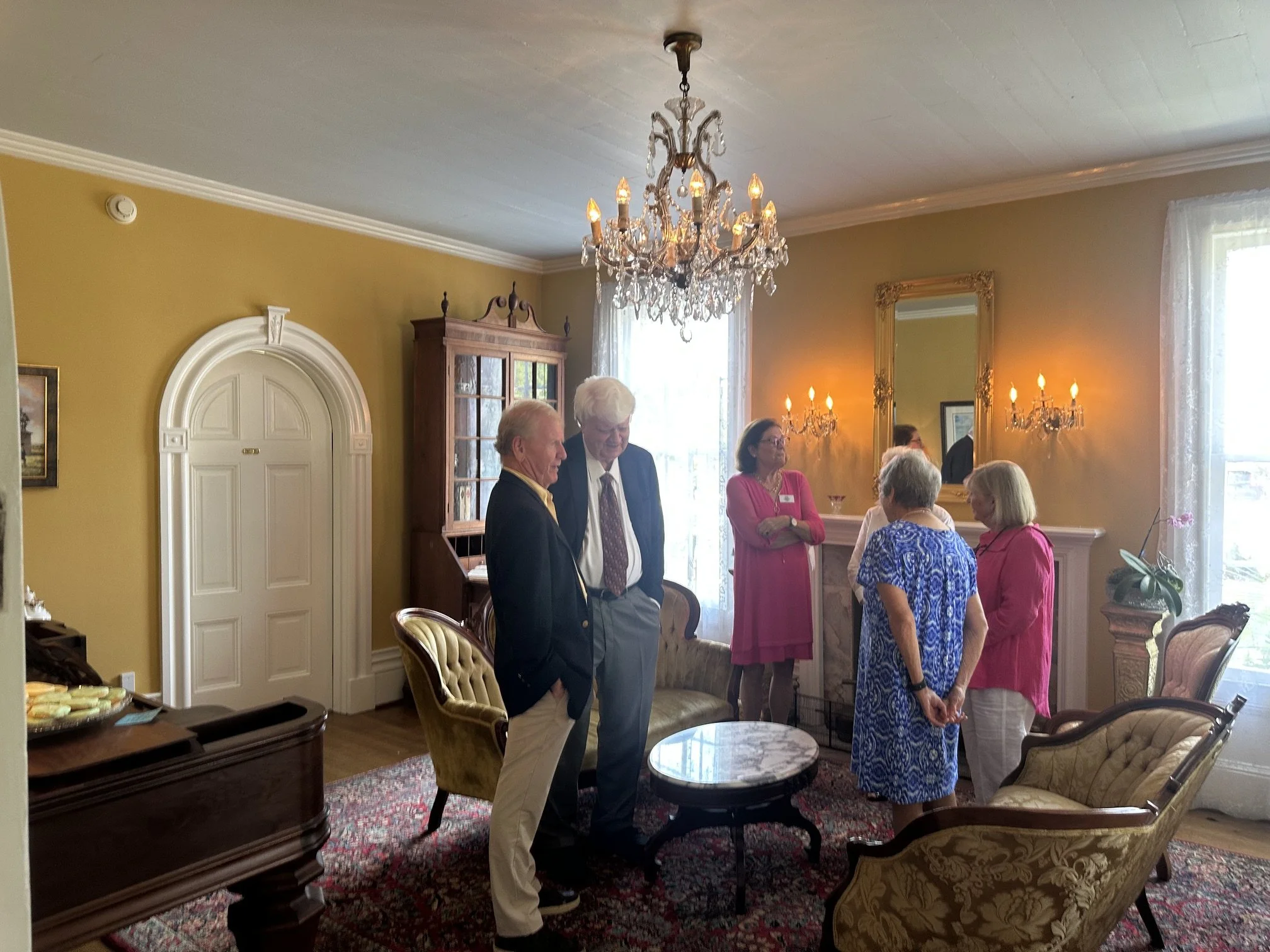 People gathered in a well-lit, elegant room with yellow walls and white crown molding, engaging in conversations. The room features a chandelier, wall-mounted sconces, a mirror, and a china cabinet. The attendees are dressed in semi-formal attire, including suits and dresses, in a social setting.