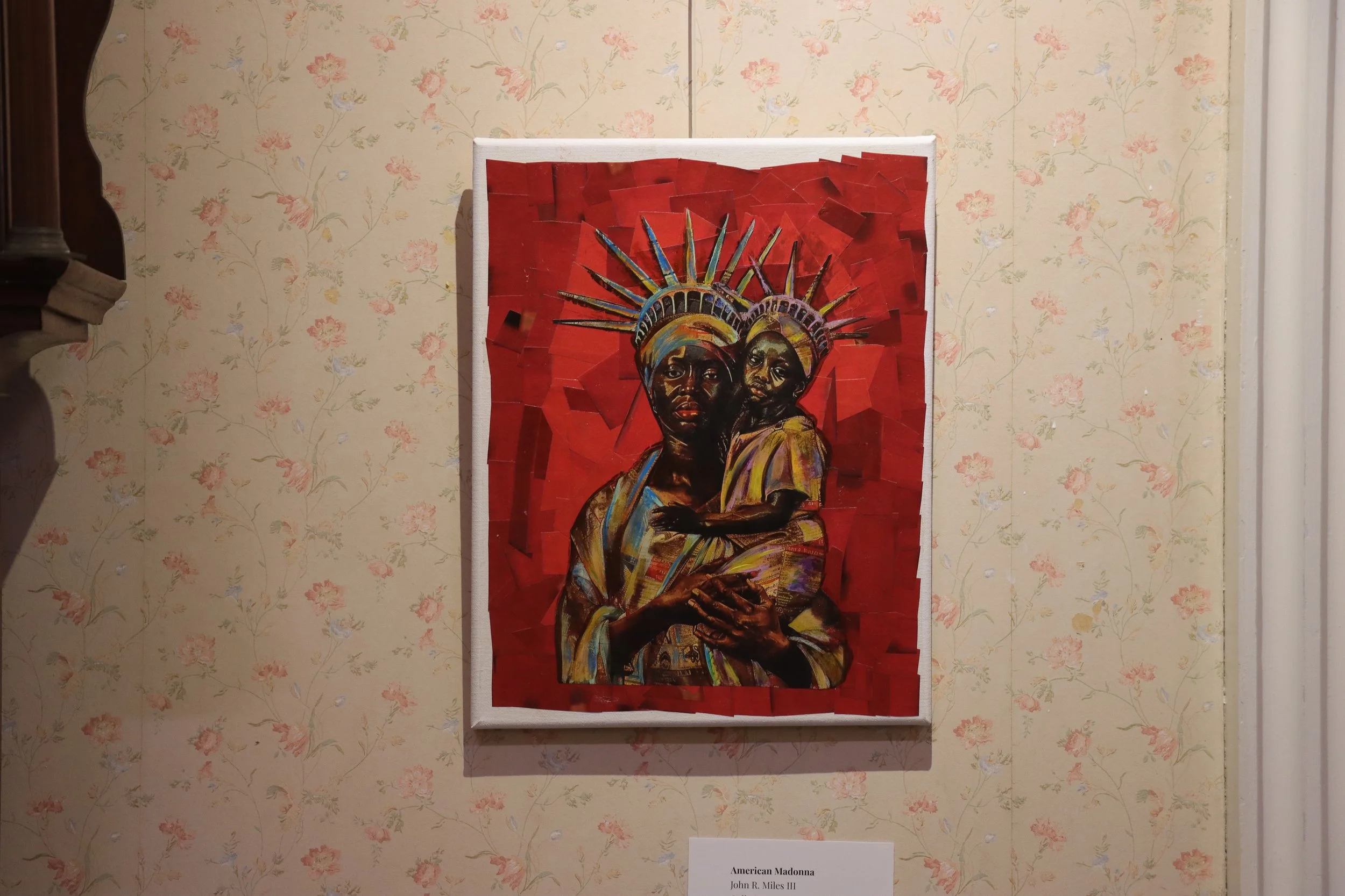 A portrait artwork depicting two African American individuals with crown-like headpieces against a red background, titled 'American Madonna' by John R. Miles III, hanging on a wall with floral wallpaper.