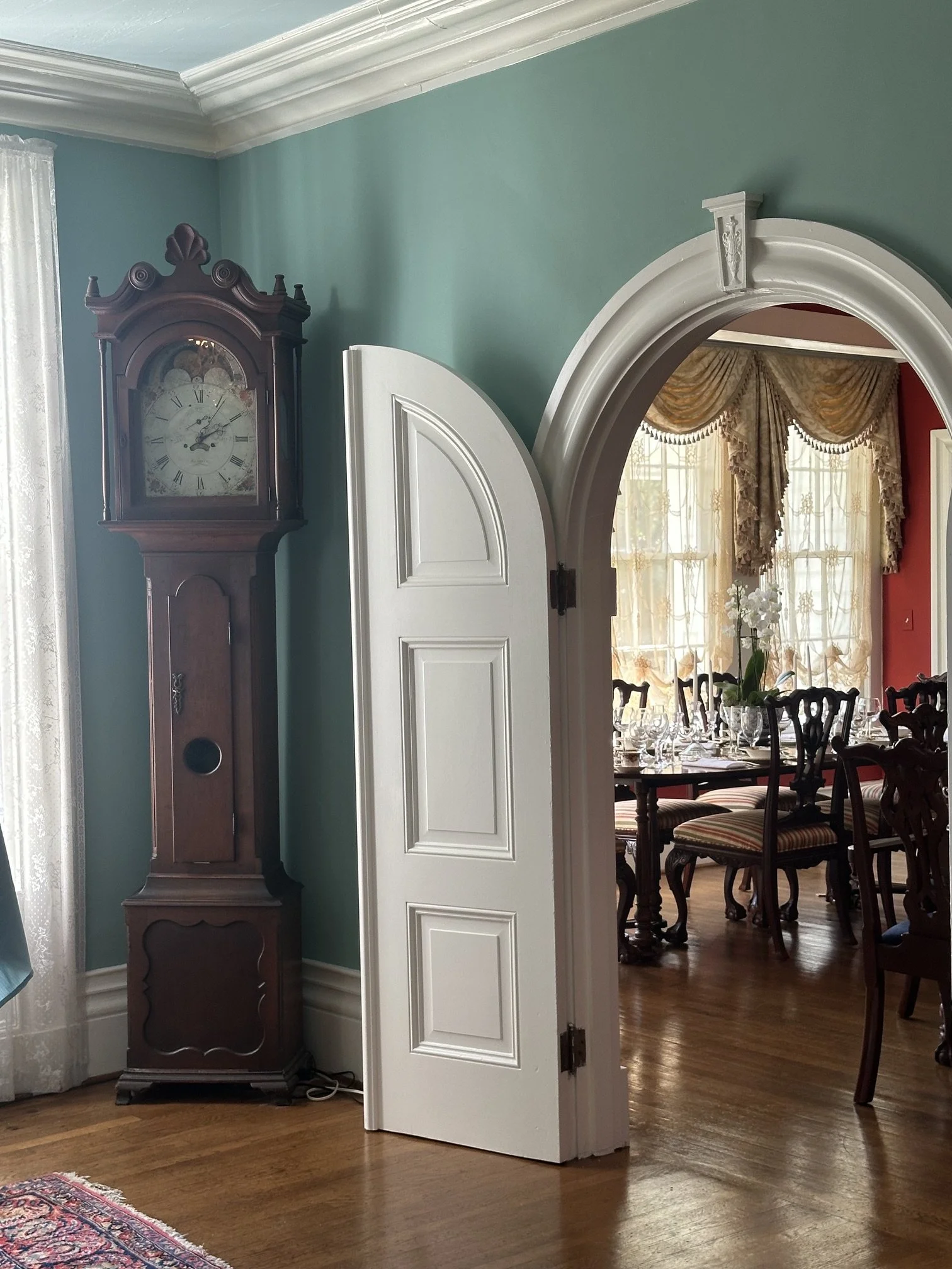 A wooden grandfather clock standing in a room with teal walls, next to a white door that is partially open, revealing a dining room with large windows, patterned drapes, and a dining table set with glassware and floral centerpiece.