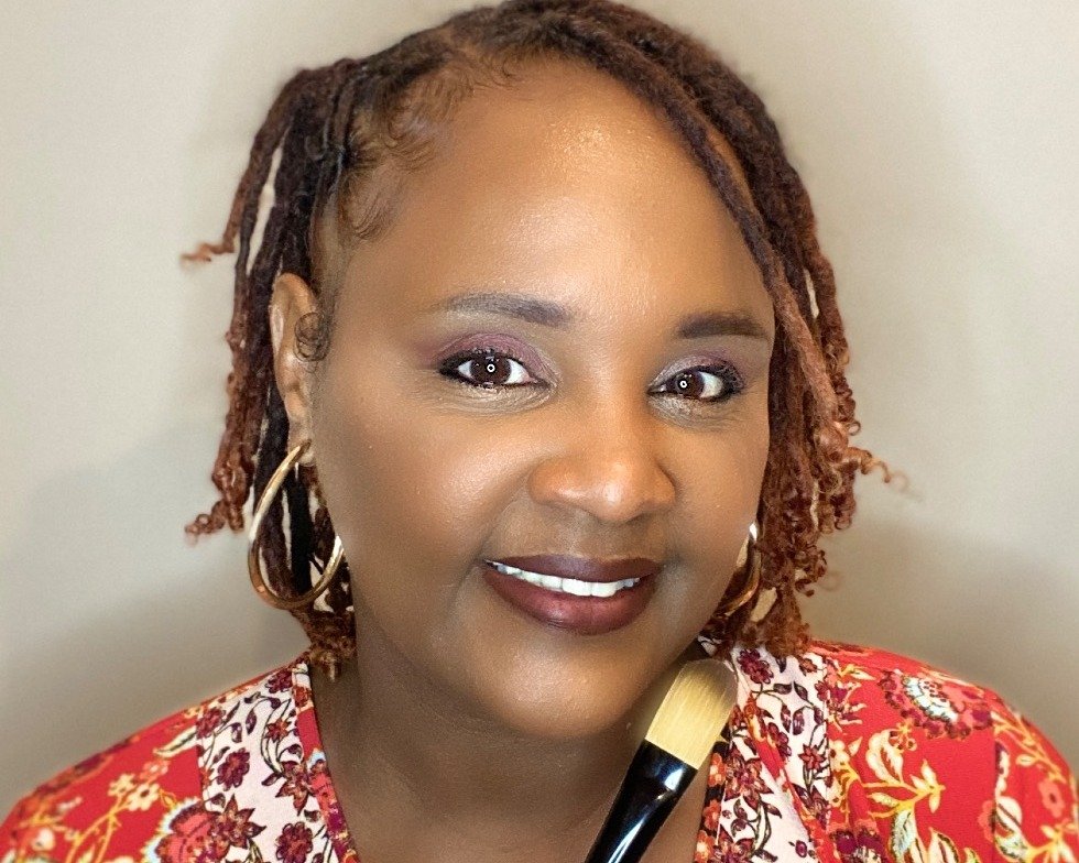 A woman with brown and red dreadlocks, wearing a colorful floral top, gold hoop earrings, and makeup with eyeshadow and lipstick, smiling at the camera, holding a makeup brush.