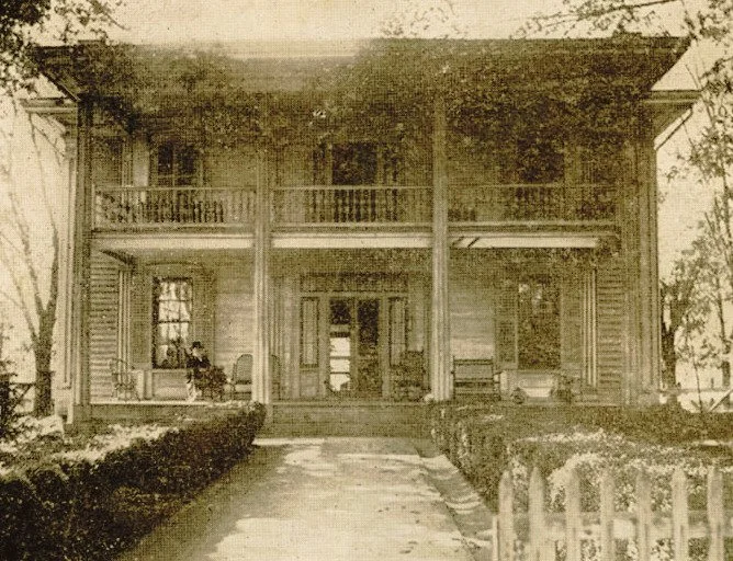 Black and white photo of a two-story house with a front porch and balcony, surrounded by trees and bushes.