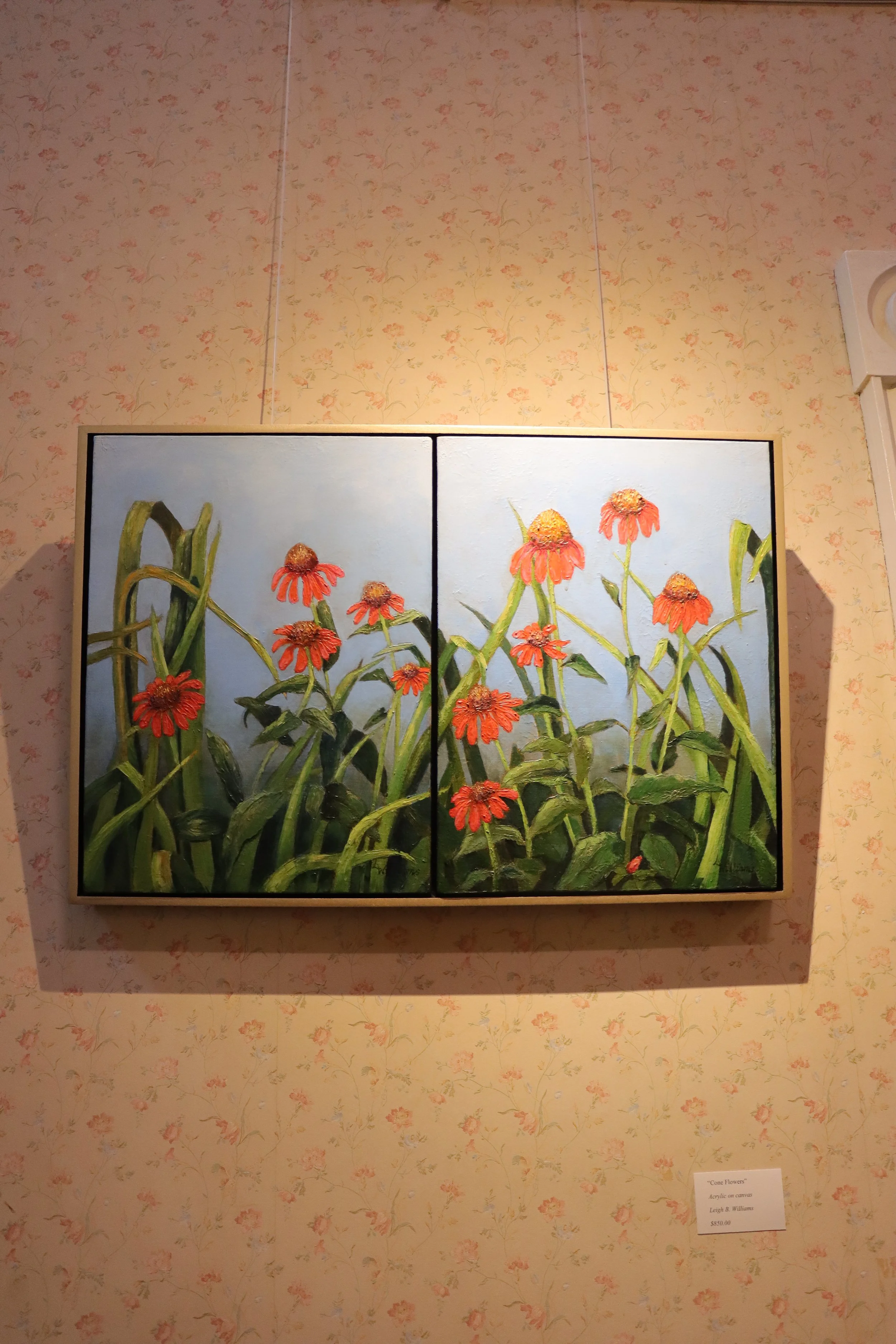 A two-panel acrylic painting of red cone flowers with green stems on a light blue background, mounted on a wall with pink floral wallpaper.