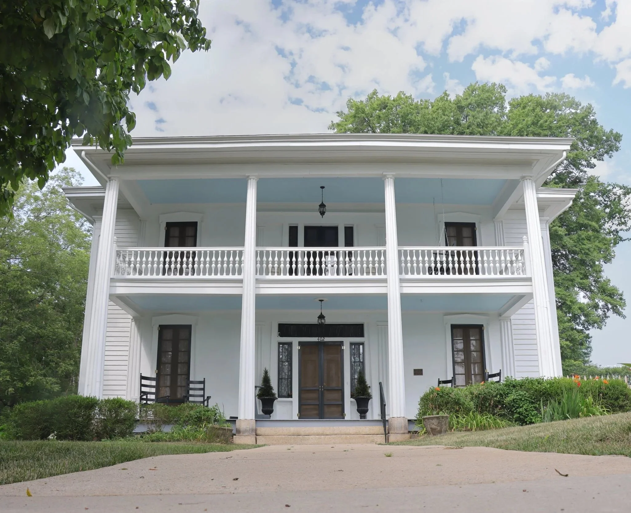 Front view of a white house with columns, a porch, black chairs, and potted plants on the porch, surrounded by greenery and trees, with a partly cloudy sky overhead.