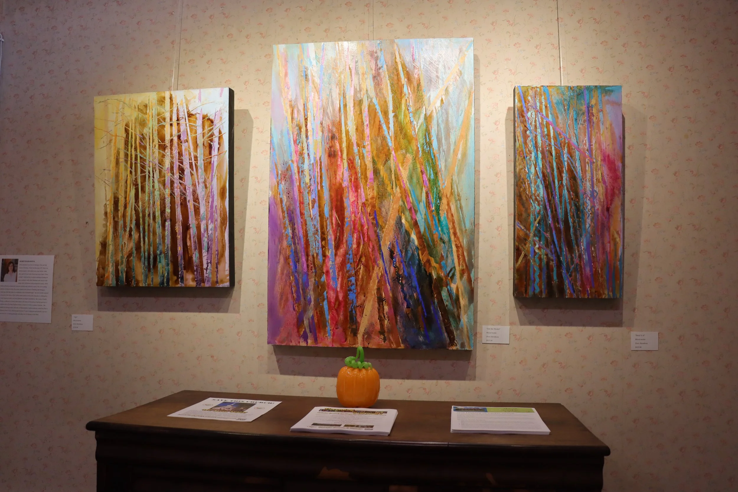 Three abstract paintings with vertical streaks of various colors hanging on a patterned wall in an art gallery, with a pumpkin decoration on a table below.