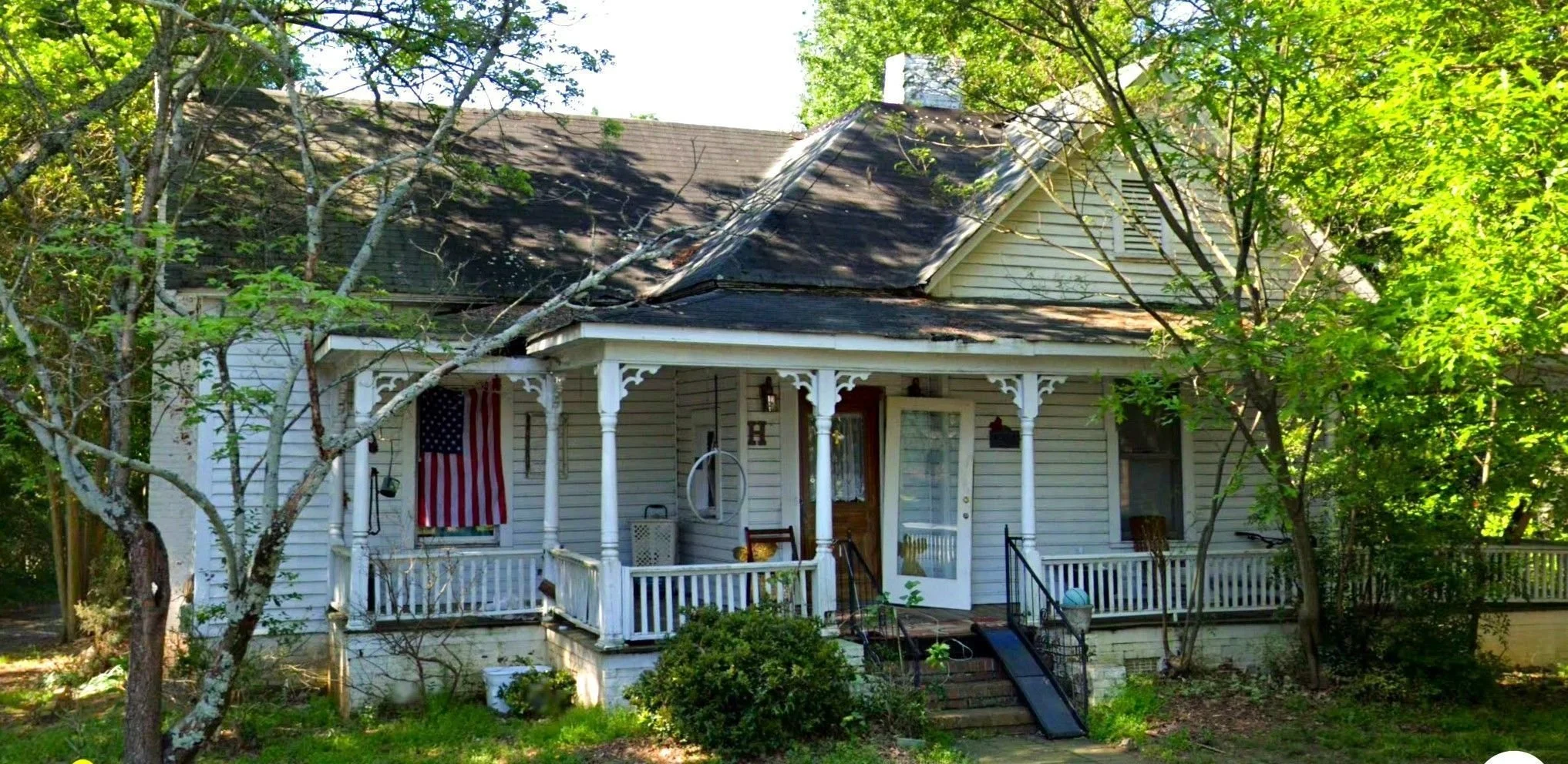 A white house with a front porch, surrounded by green trees and shrubs, with an American flag hanging on the porch.