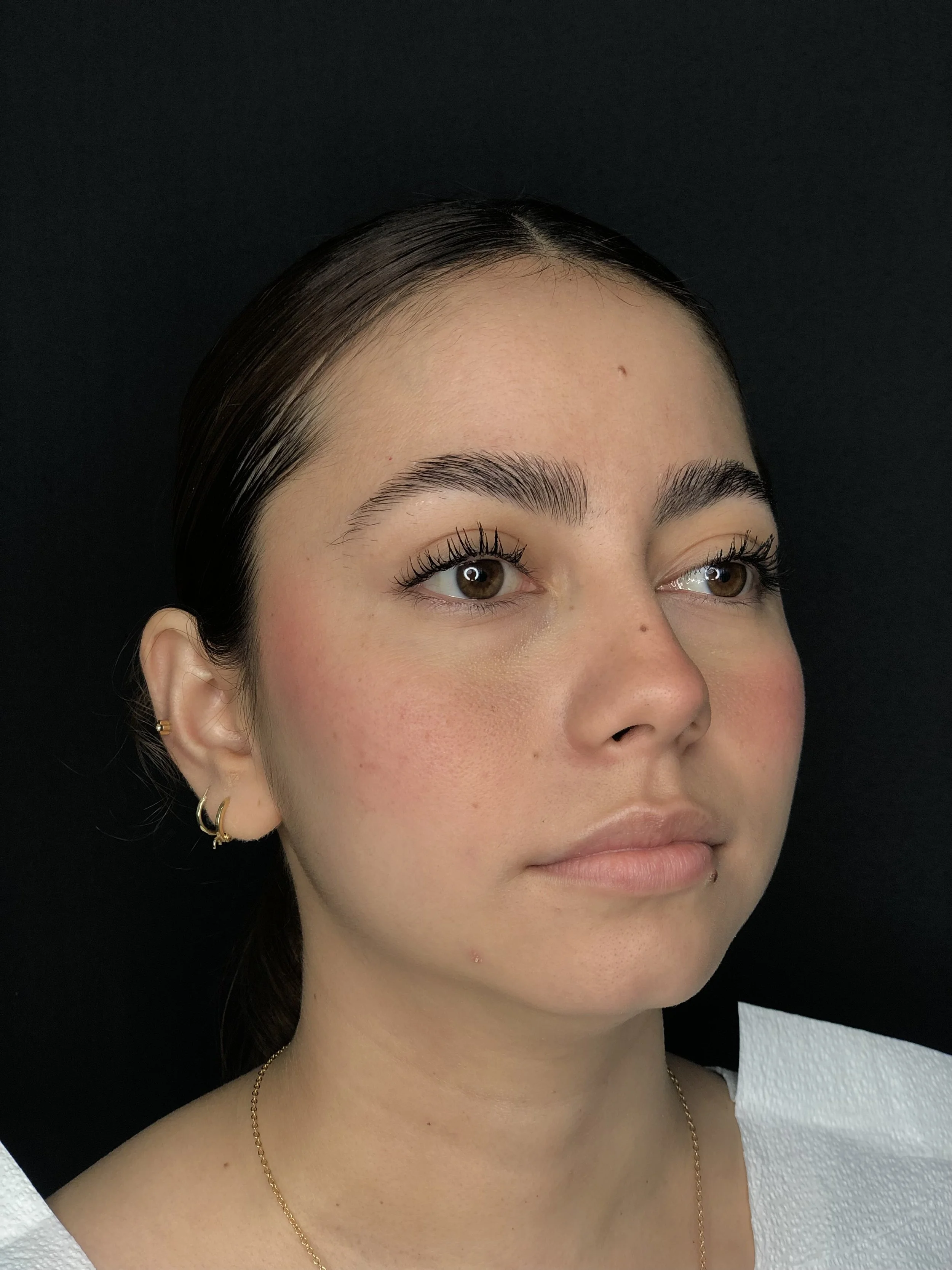 Close-up of a young woman's face with makeup, dark hair, and earrings, against a black background.