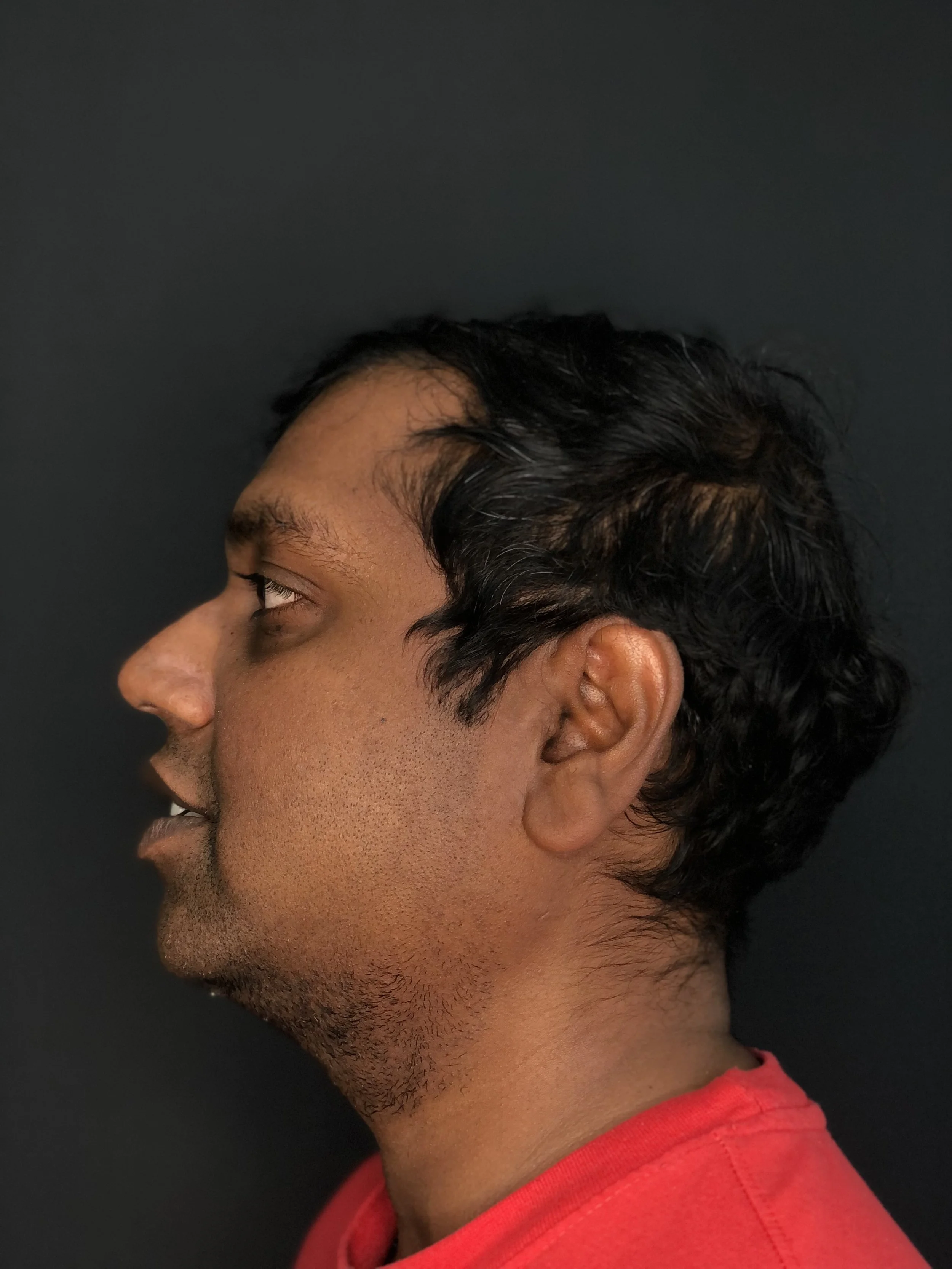 Side profile of a man with dark hair and facial hair, wearing a red shirt against a black background.