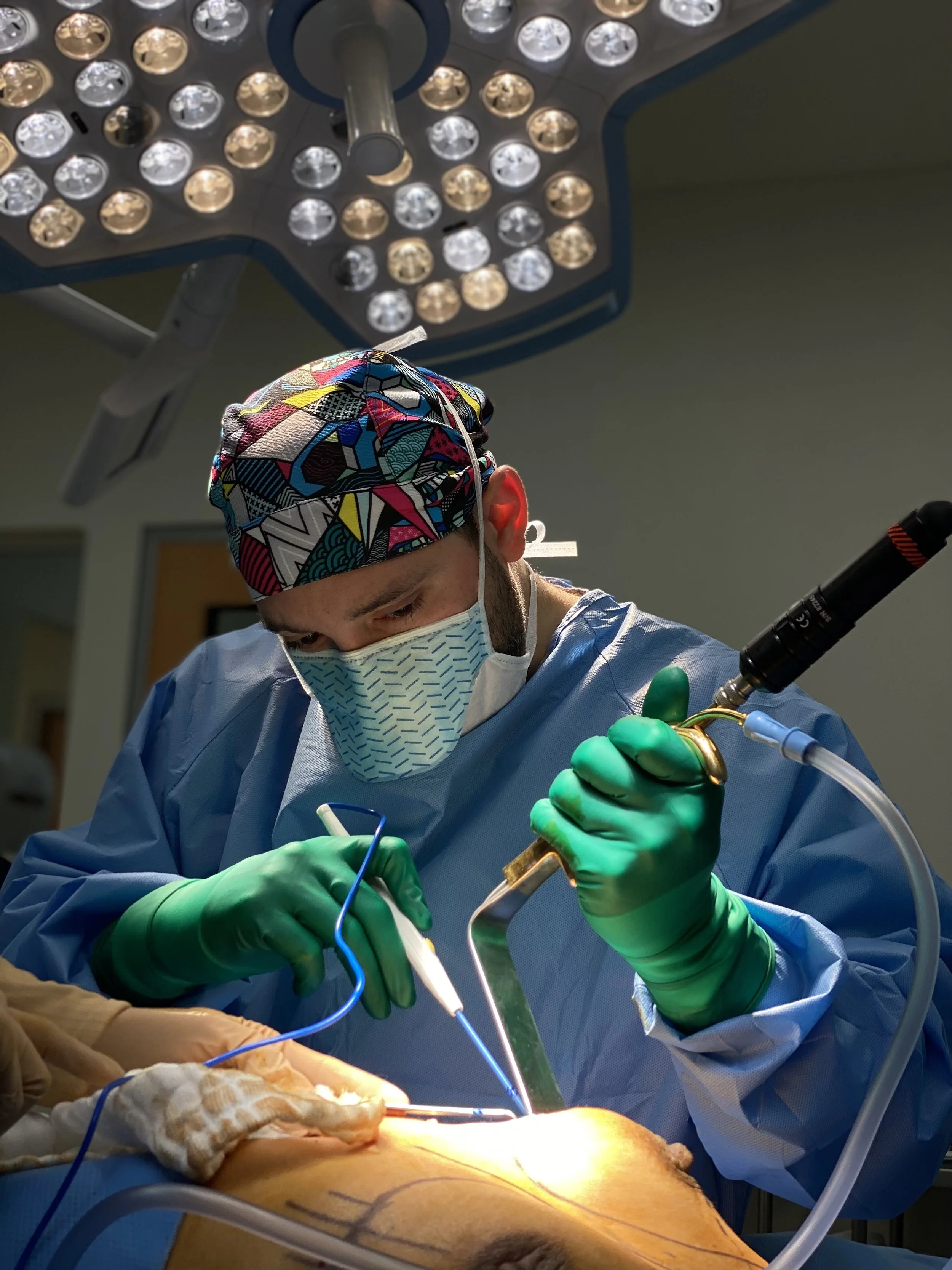 Surgeon performing surgery in an operating room, wearing a colorful scrub cap, face mask, and green gloves, with surgical tools and bright lights overhead.