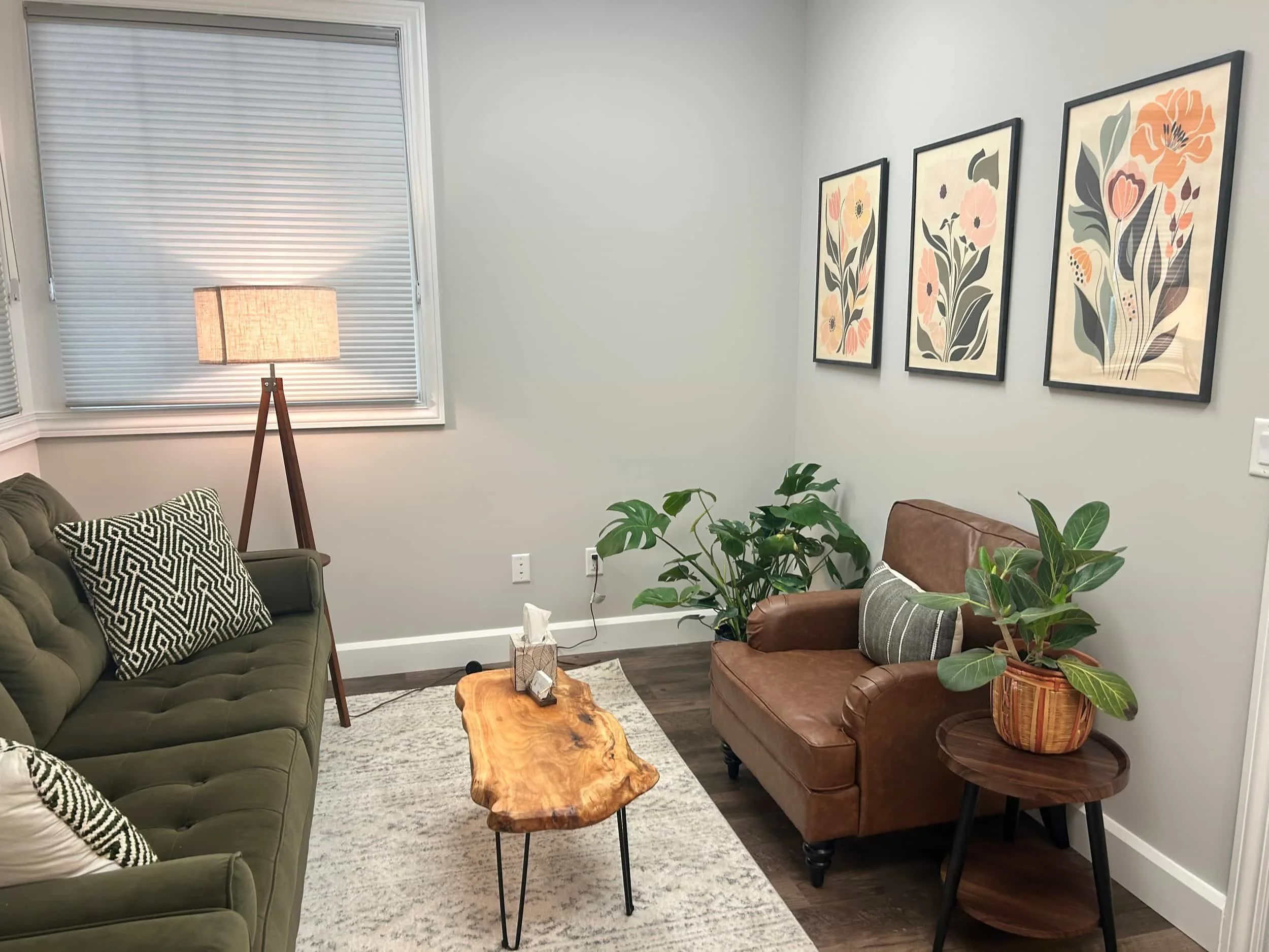 A cozy living room with a green sofa, brown armchair, wood coffee table, plants, and floral art on light gray walls.