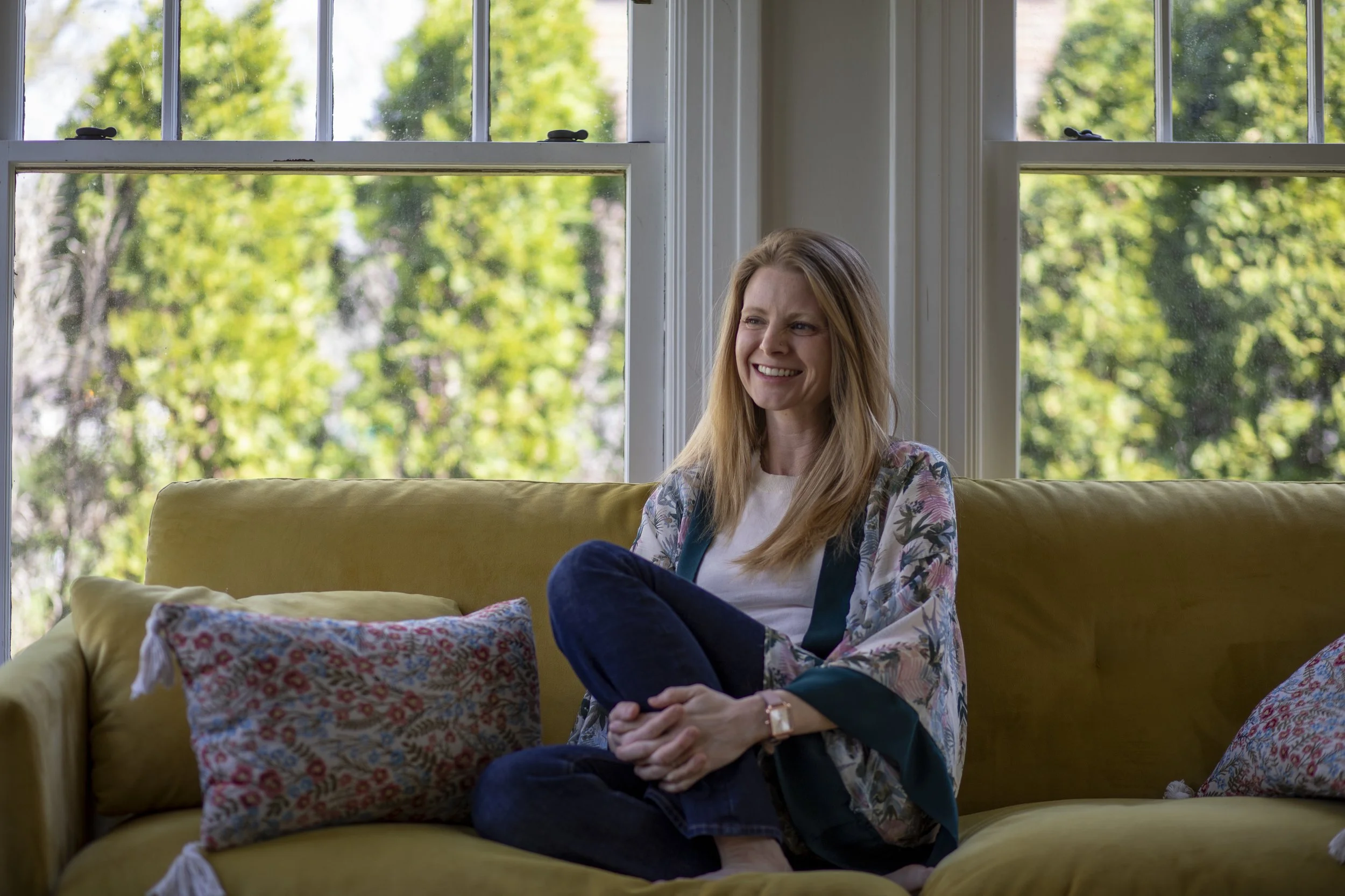 A woman with long hair sits cross-legged on a yellow couch, smiling, with large windows and green trees in the background.
