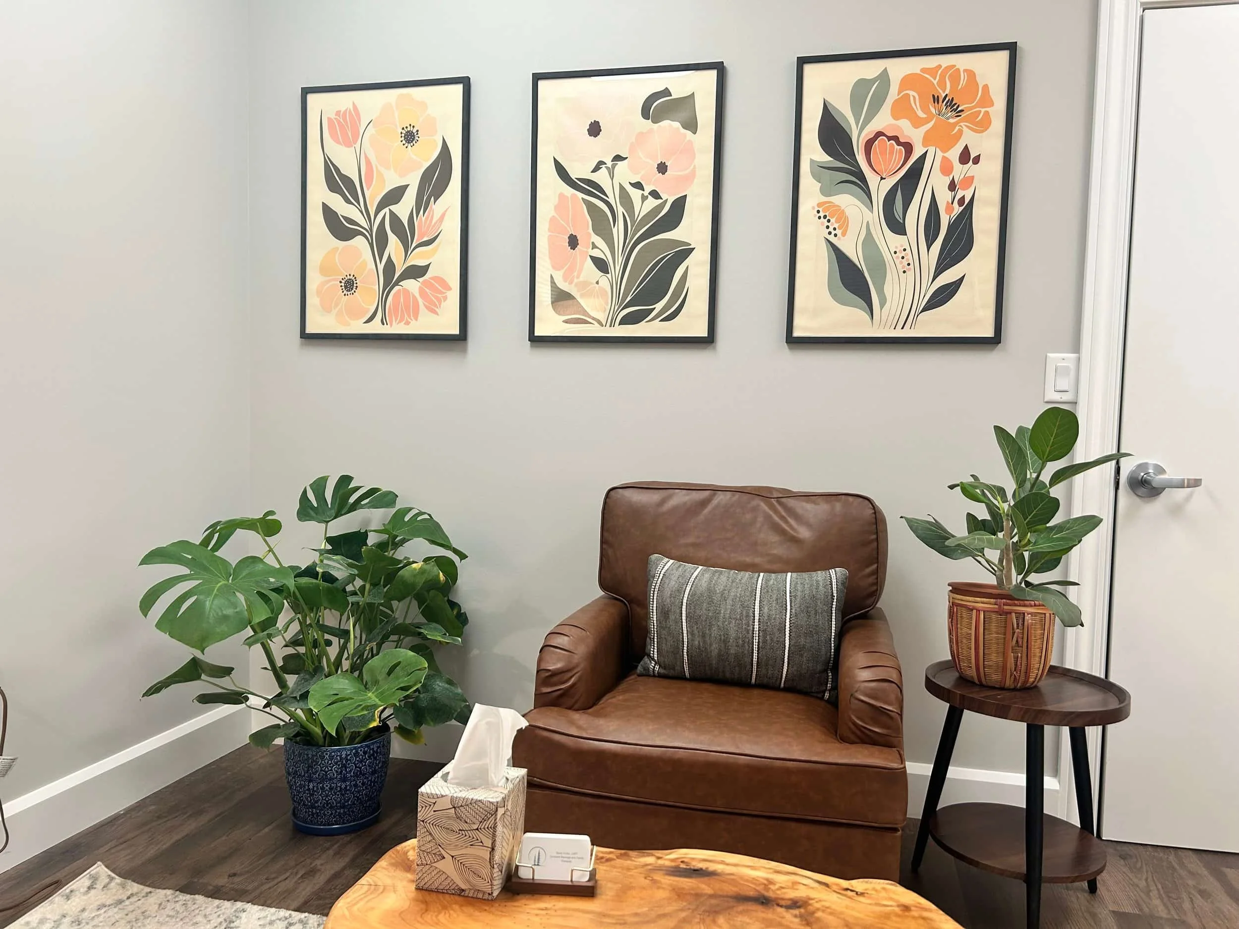 A cozy room with a brown leather chair, green plants, a side table, and three floral art prints on a light gray wall.