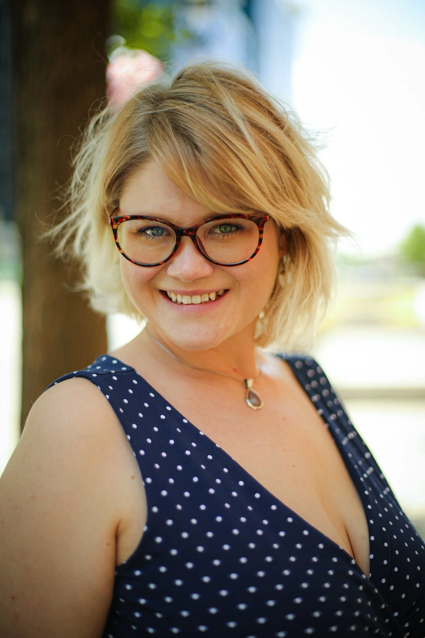 A woman with blonde hair, wearing glasses, a navy blue dress with white polka dots, and a necklace, smiling outdoors with a blurred green background.