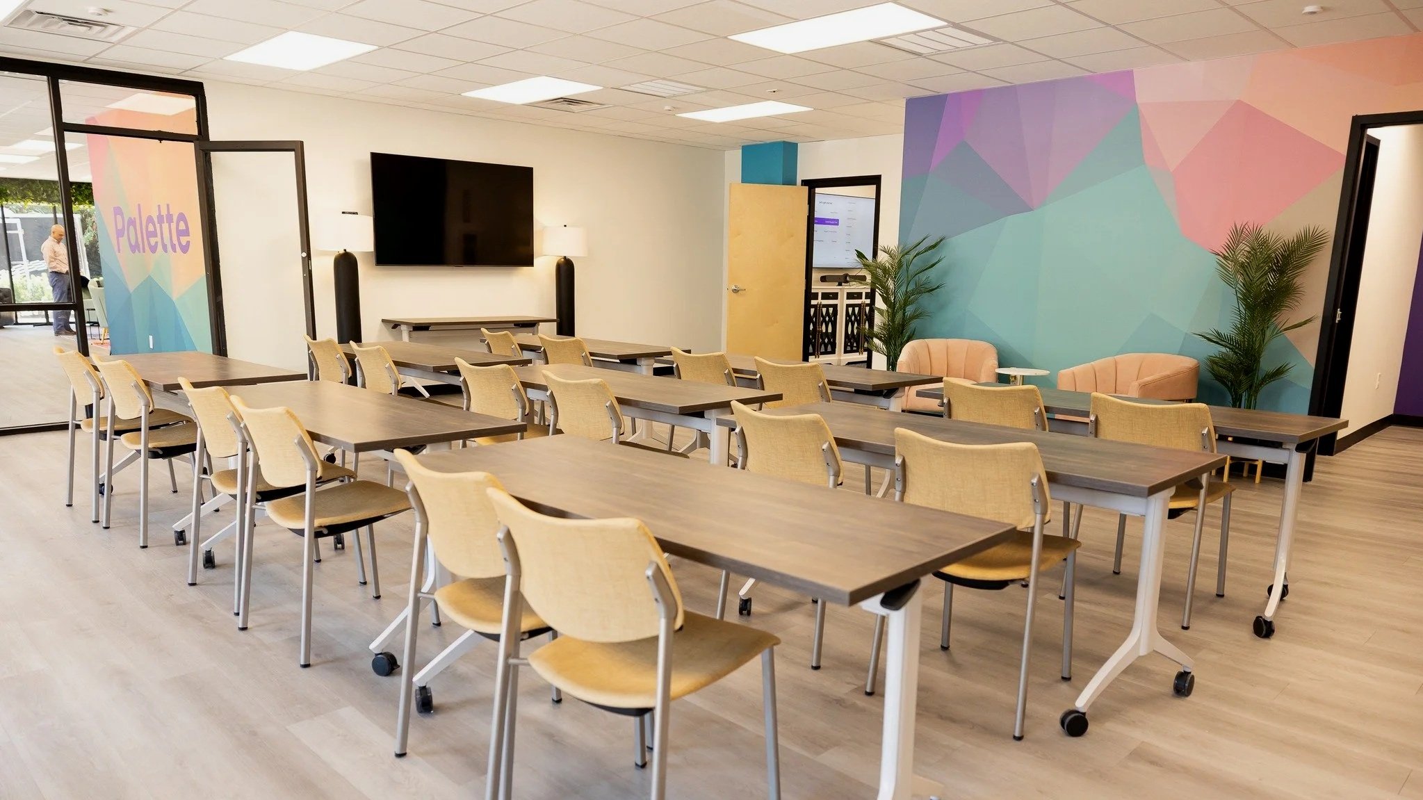 Meeting room with wooden tables, beige chairs, a pink armchair and small table, large wall mural with pastel geometric shapes, plants, and a wall-mounted TV.