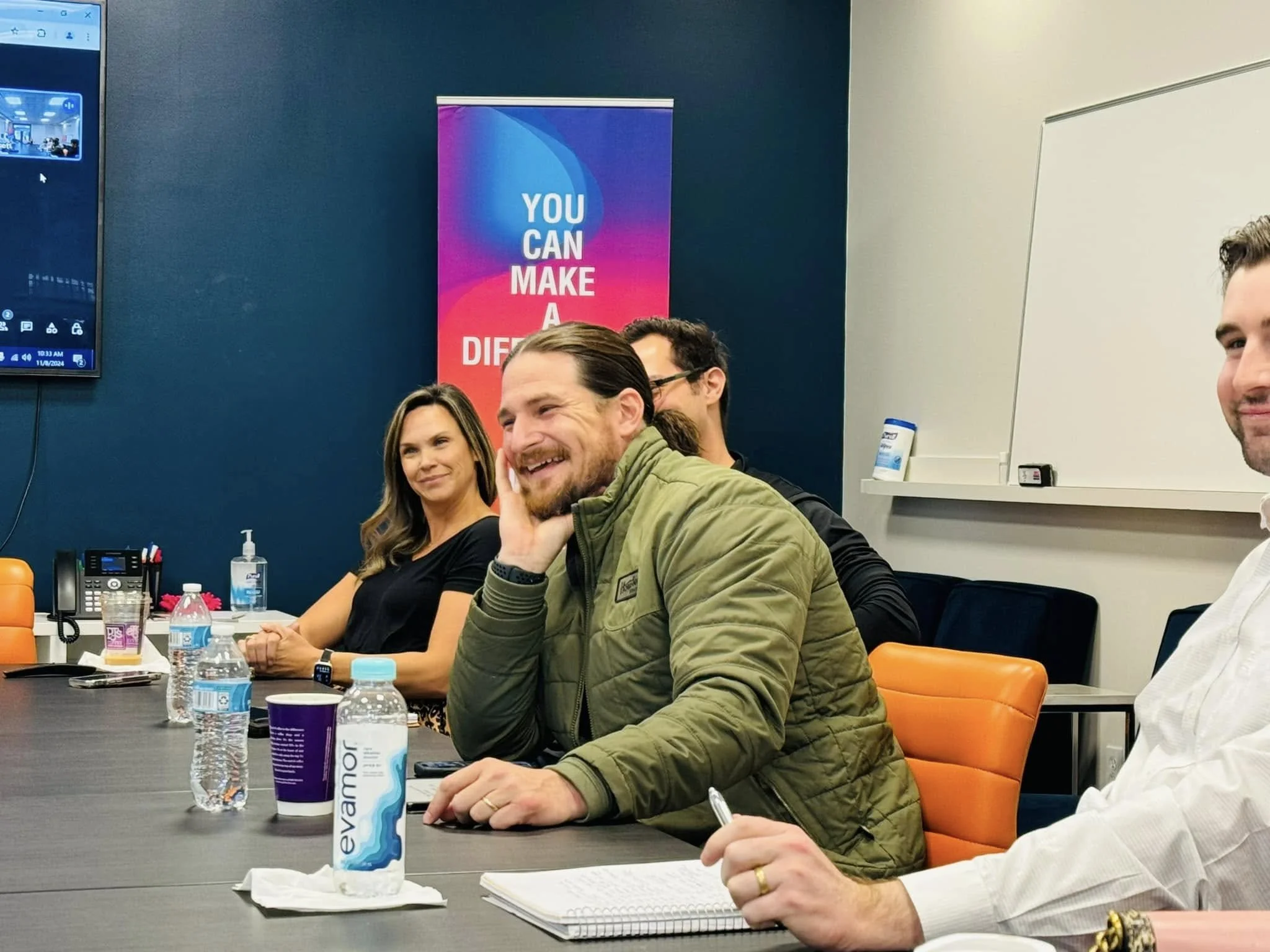 People sitting around a conference table during a meeting, with water bottles, paper cups, notebooks, and writing utensils on the table. A large Dell monitor is mounted on the wall displaying a digital interface. A motivational poster with the text "You Can Make a Difference" is visible in the background.