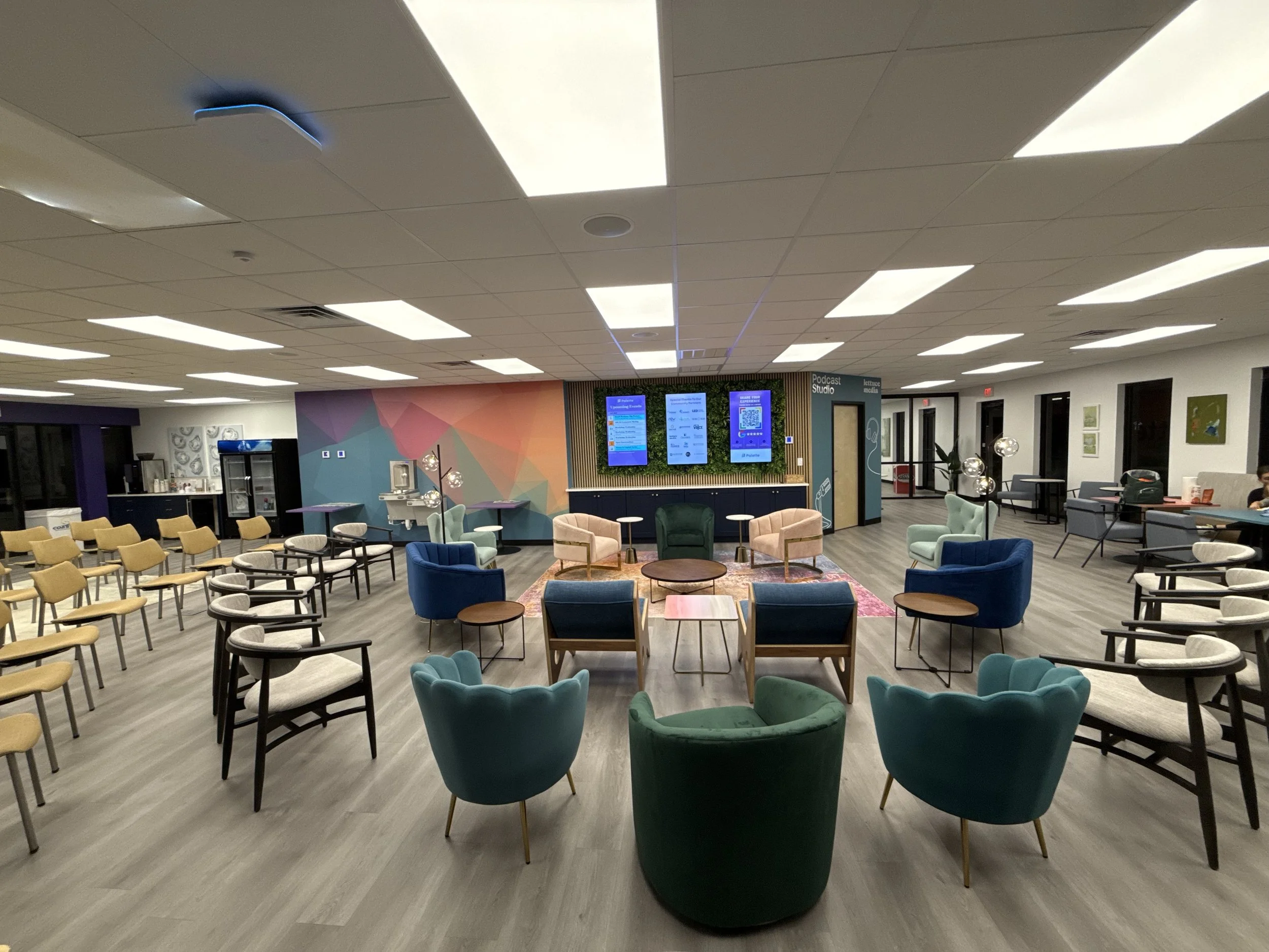 An interior view of a modern, well-lit lounge or waiting area with a variety of chairs and tables, a colorful geometric wall, digital screens, and decor such as plants and artwork.