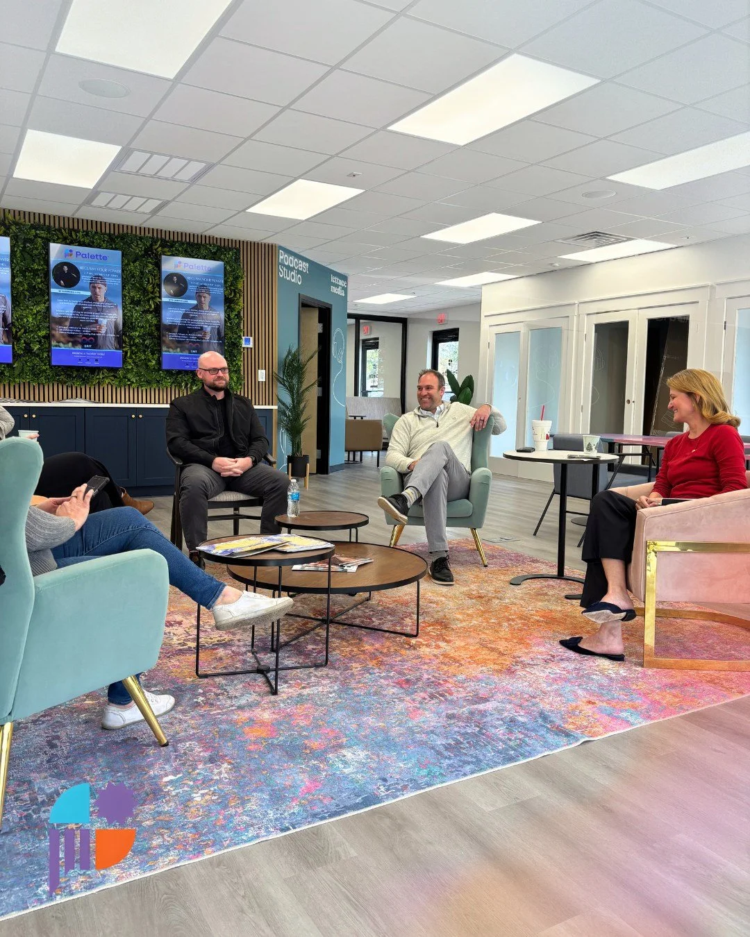 People sitting and conversing in a modern, colorful office lounge with a vibrant patterned rug and wall-mounted screens.
