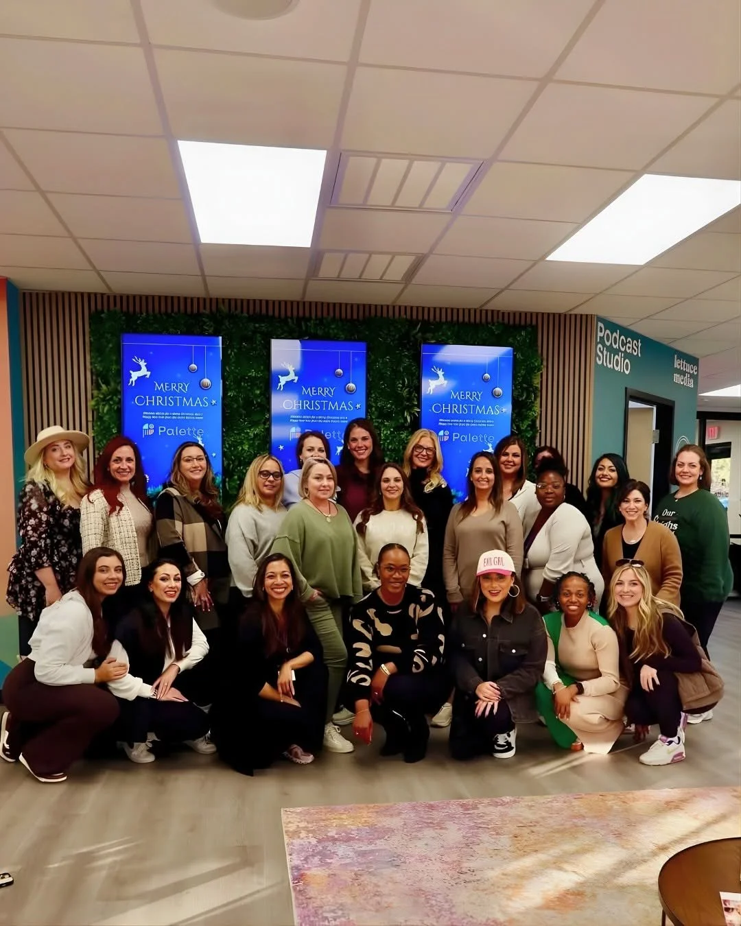 Group of women gathered in an indoor space, celebrating Christmas with digital screens displaying holiday greetings in the background, and a sign indicating a podcast studio nearby.