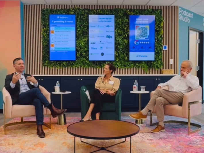 Three people sitting in a panel discussion in a modern conference room with a green wall and digital screens behind them. Two men and one woman, each with a water bottle on a small table beside them.