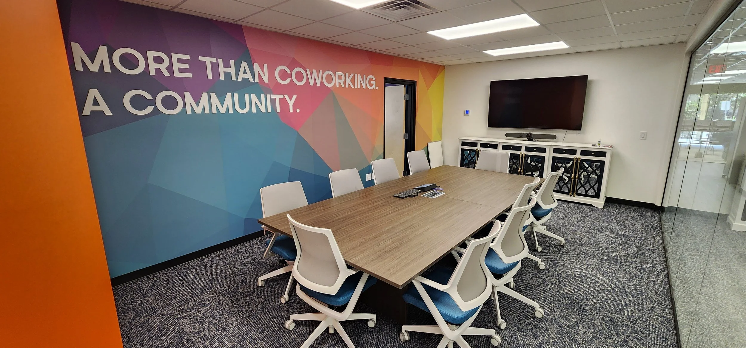 A conference room with a wooden table surrounded by white office chairs with blue cushions. The wall has a colorful geometric mural with the words 'More Than Coworking. A Community.' In the room, there is a large flat-screen TV, a white cabinet, and a glass wall.