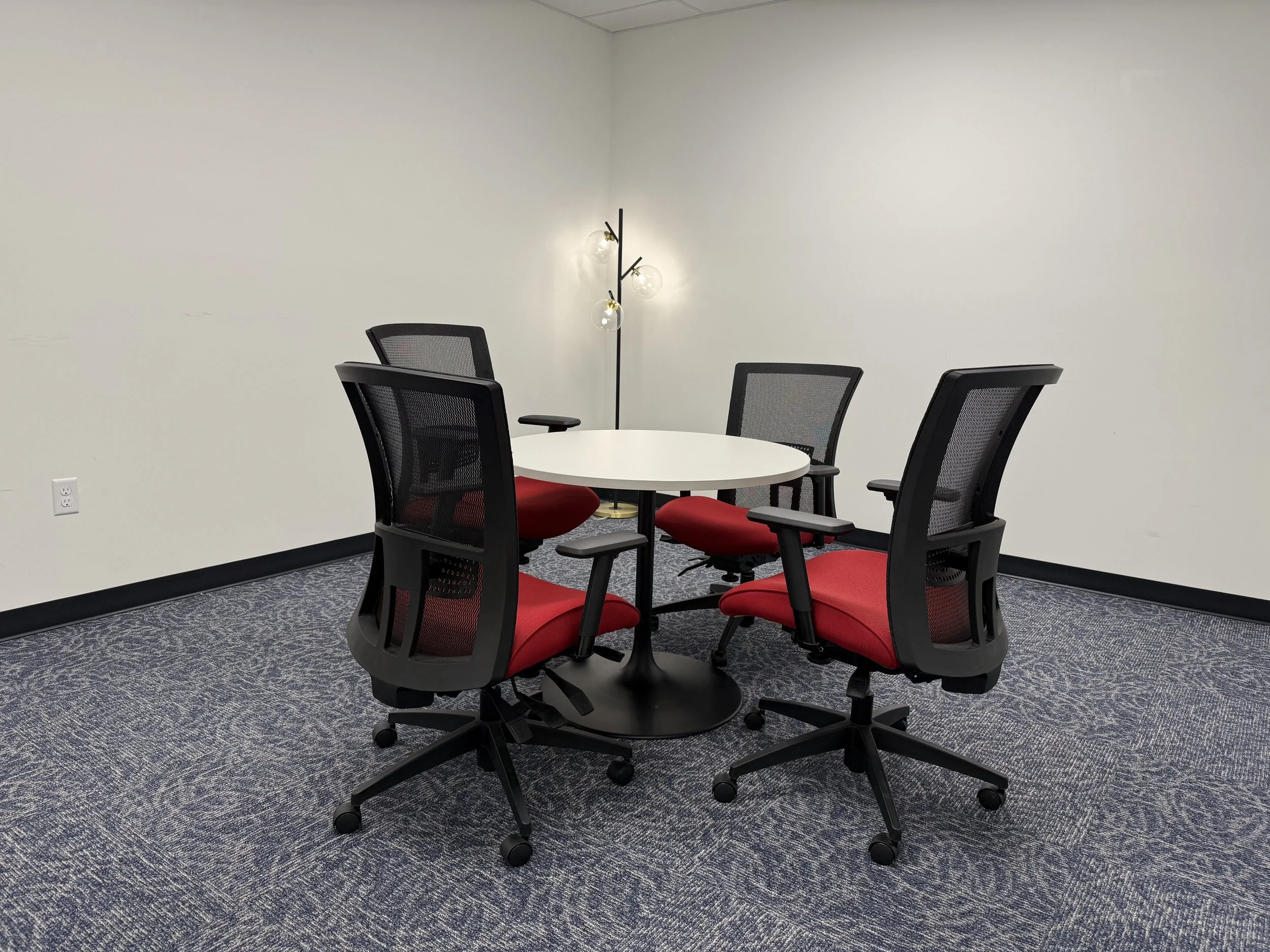 A modern conference room with a round white table surrounded by six black office chairs with red cushions and mesh backrests. In the corner, there is a black floor lamp with three glass globe bulbs. The walls are plain white, and the floor is covered with a blue patterned carpet.