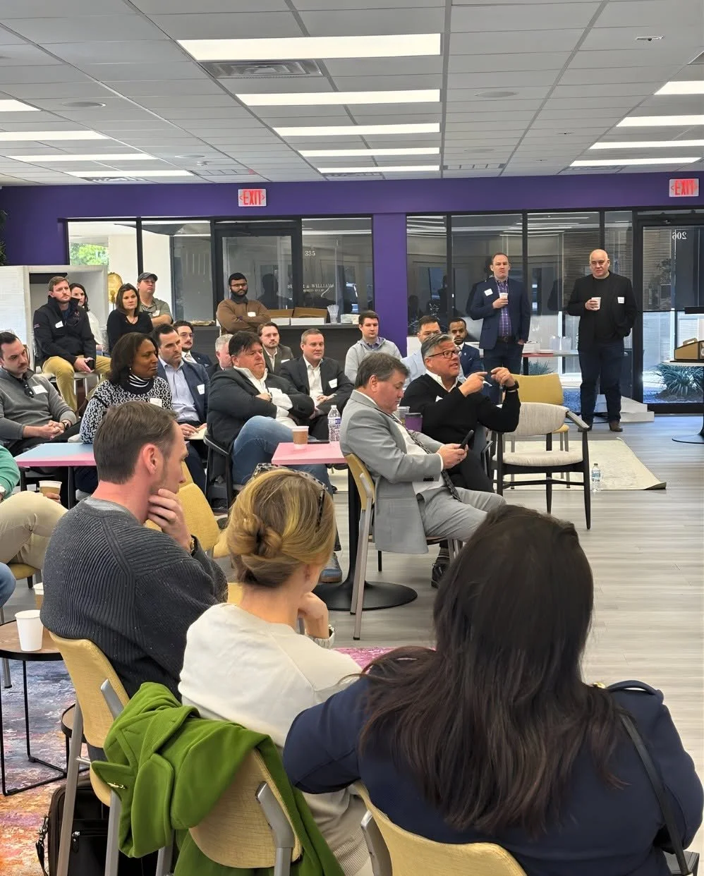 People attending a professional event or seminar in a modern office space, sitting and listening to speakers at the front, some taking notes or using devices, with a purple accent wall and glass doors in the background.