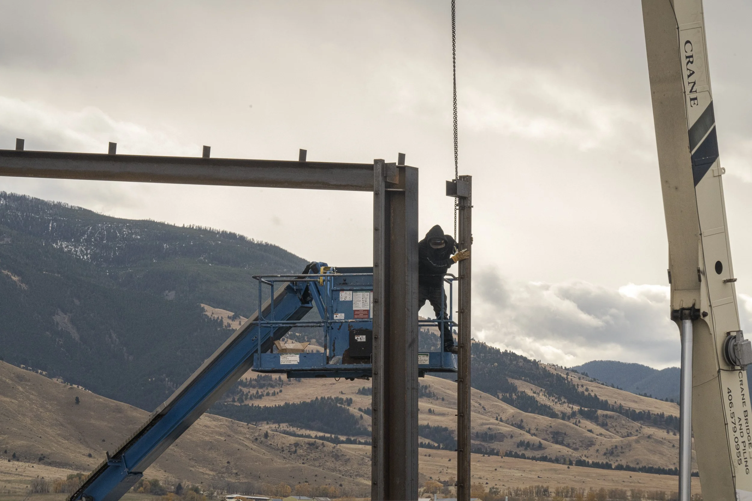 A steel worker erecting structural steel on a house near bozeman montana. The clouds fill the sky with small patches of snow in the trees.