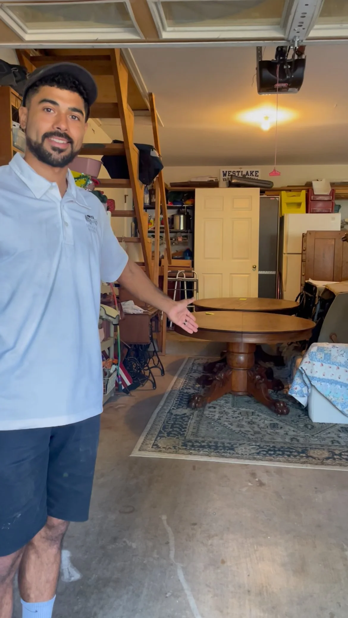 Man in white polo shirt and black shorts standing in cluttered garage, pointing at a round wooden dining table with a dark stained finish and ornate claw feet, with a patterned area rug underneath.