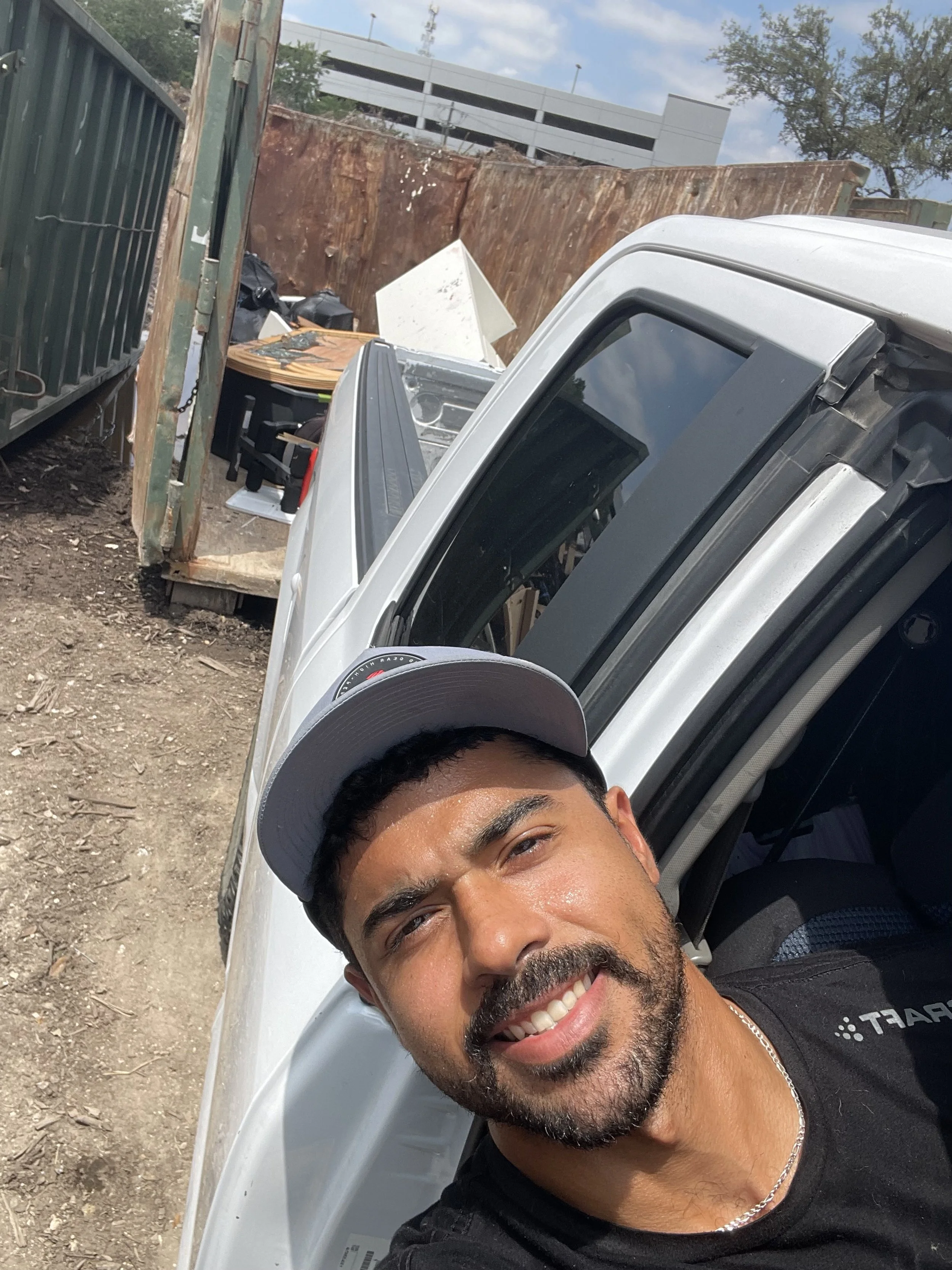 alt="A man taking a selfie leaning against a white vehicle with a smile. Behind him is a dusty area with a rusted dumpster, a white piece of furniture, and some outdoor furniture, under a partly cloudy sky".