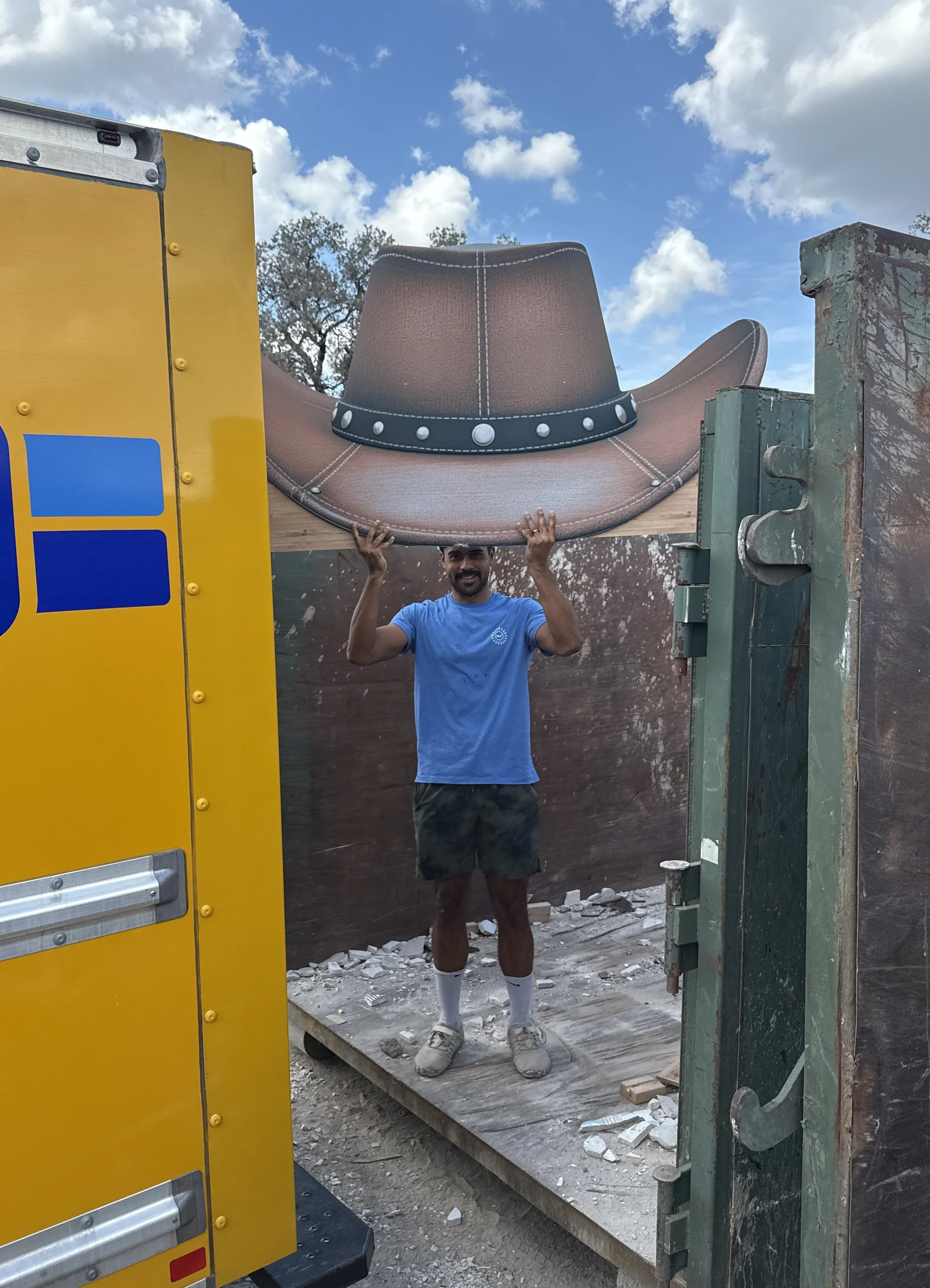 A man standing outdoors, holding a large, cowboy hat-shaped sign above his head with both hands, smiling. The sky is partly cloudy and there are construction materials on the ground.