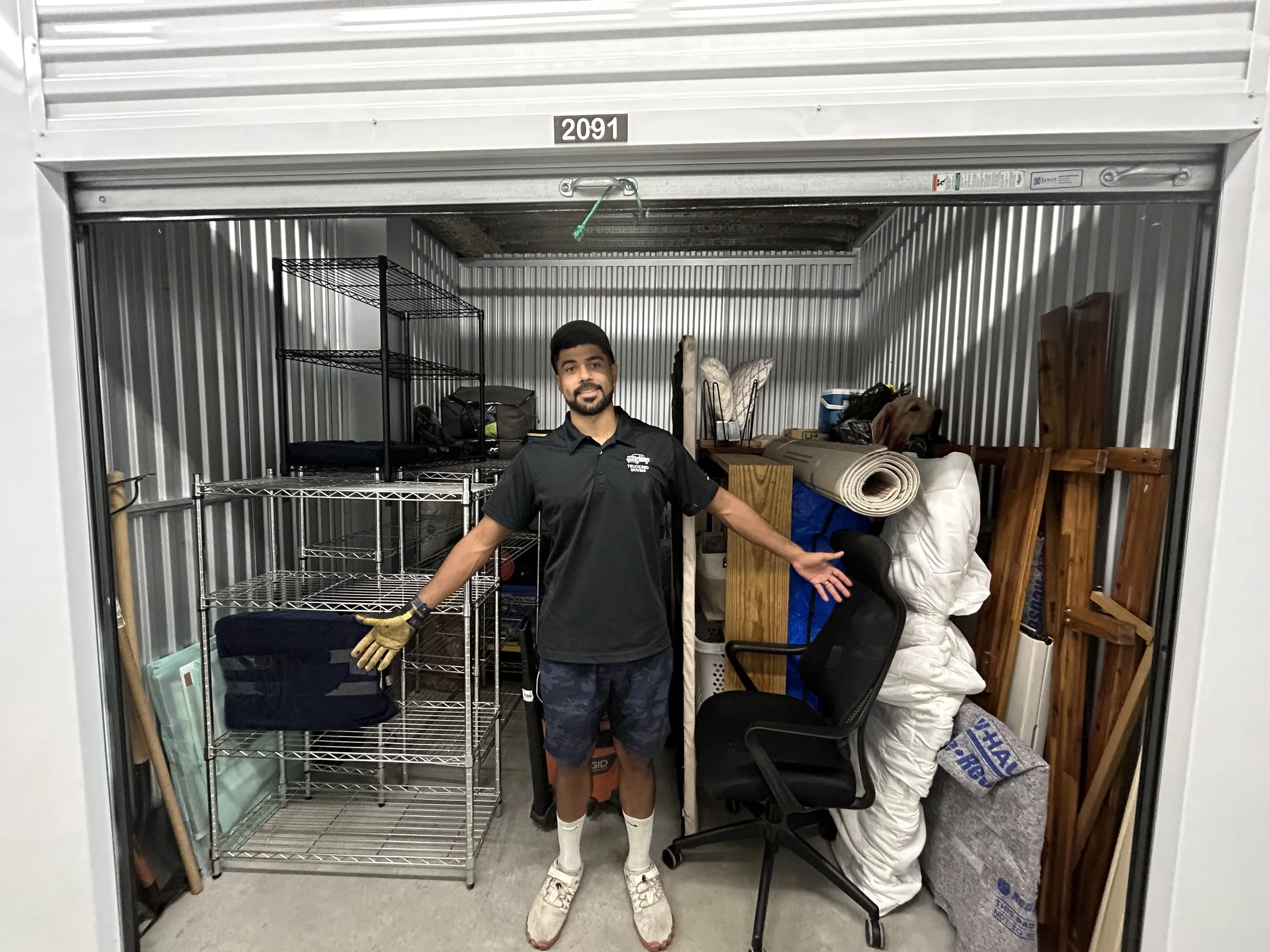 Man in black shirt and shorts standing inside a storage unit with metal shelving, wooden furniture, rolled-up carpets, and packed items.