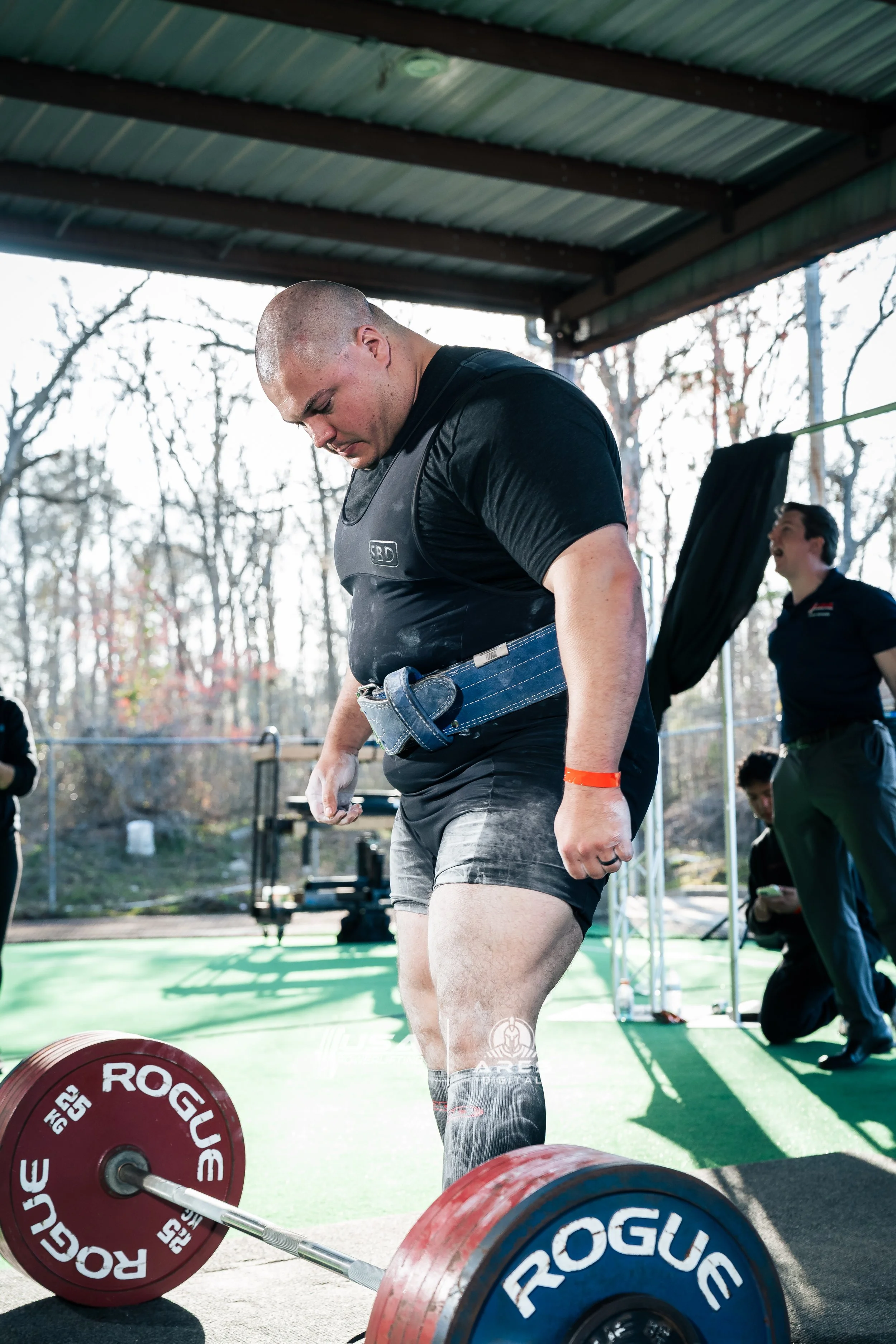 A young man in athletic gear stands on a green outdoor gym floor, preparing for lift weights, with a barbell with red and blue weight plates on the ground in front of him. He is wearing a black shirt, gray shorts, and knee-high socks, with a weightlifting belt around his waist. There are people in the background, some smiling, and trees and a roof overhead.