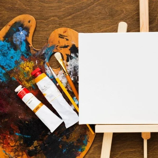 A small white canvas and light wood easel, on top of a dark wood table, next to a used wood paint palette, a couple small tubes of paint, and two paintbrushes.