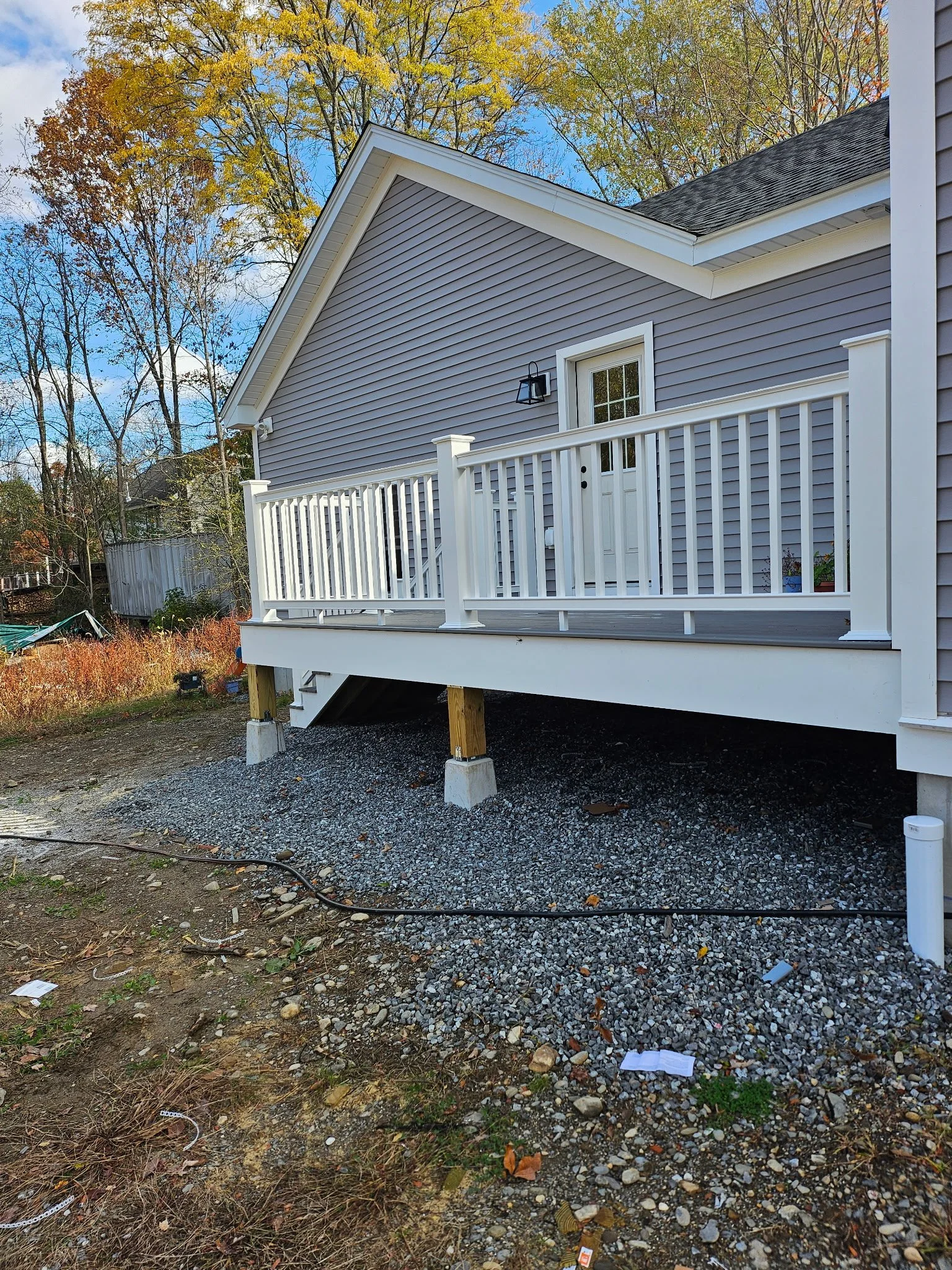 Exterior view of a house with purple siding, a white deck, and white railing, with a door, a outdoor light fixture, and a landscape with trees and a blue sky.
