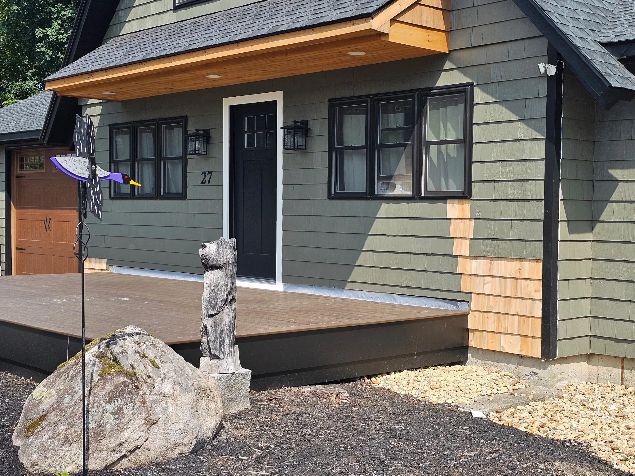 Newly constructed house with green siding, black window frames, black front door, wooden porch, and a decorative weather vane shaped like a bird on a cone-shaped base, situated near a large rock and a wooden sculpture.