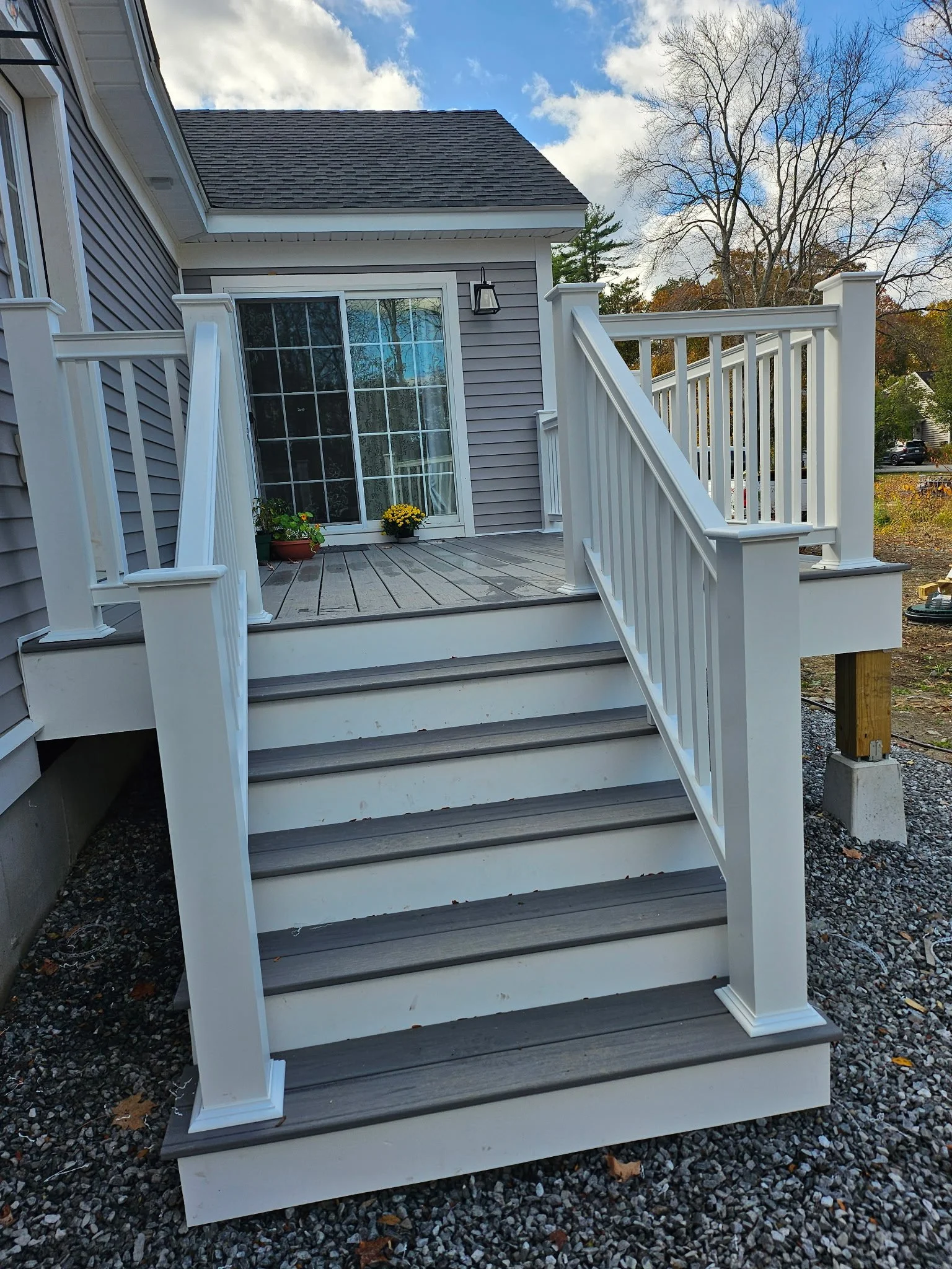View of a newly installed white wooden porch staircase leading to a back door of a house with gray siding. The porch has a railing on both sides, and the stairs are painted white with gray treads. There are two potted yellow flowers on the porch near the door. The house has a sliding glass door, and a black outdoor wall lantern is mounted above it. The background features trees with fall foliage and partly cloudy sky.