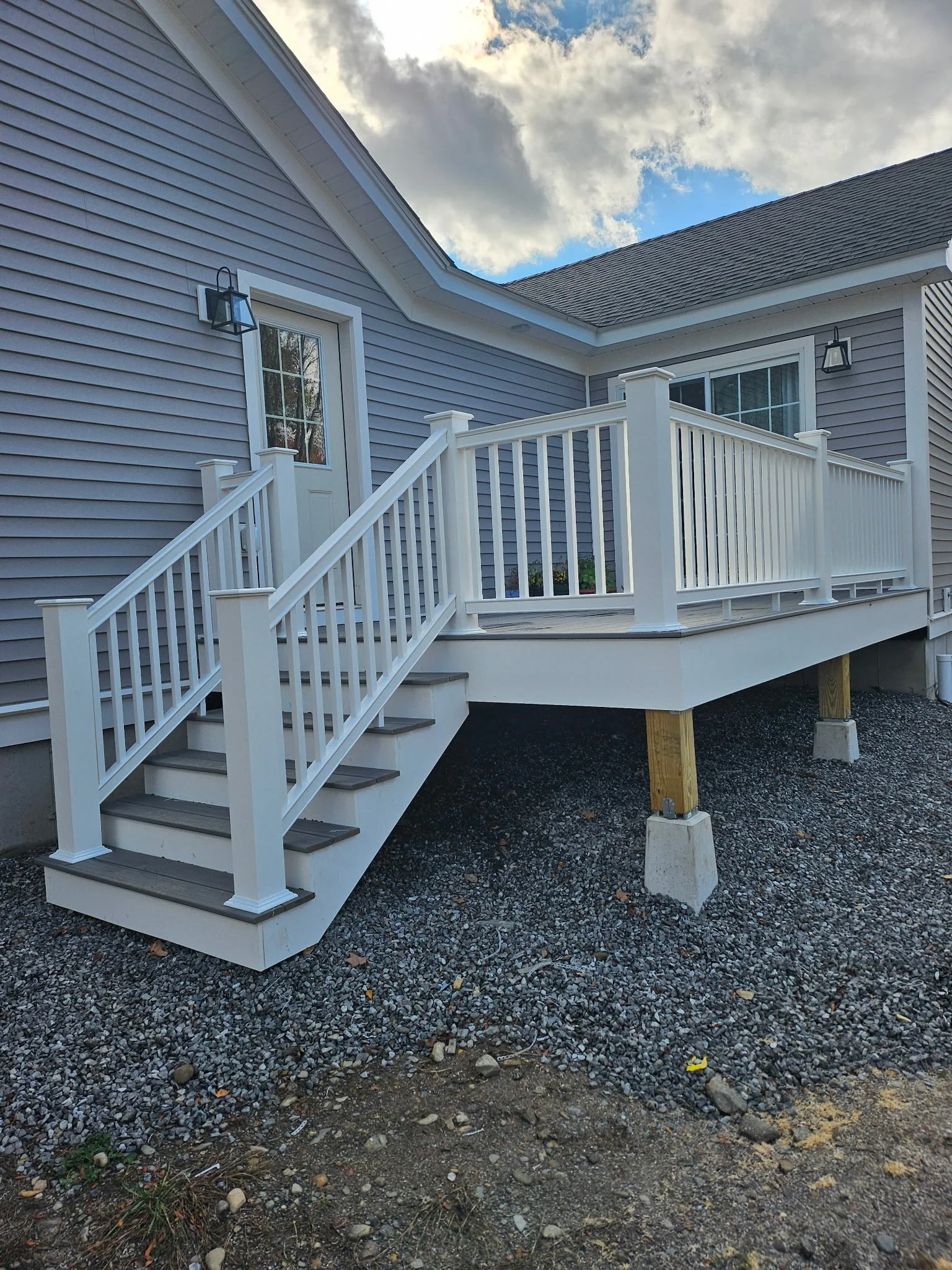 White wooden deck with stairs and railing attached to a gray house, with outdoor lighting and cloudy sky above.