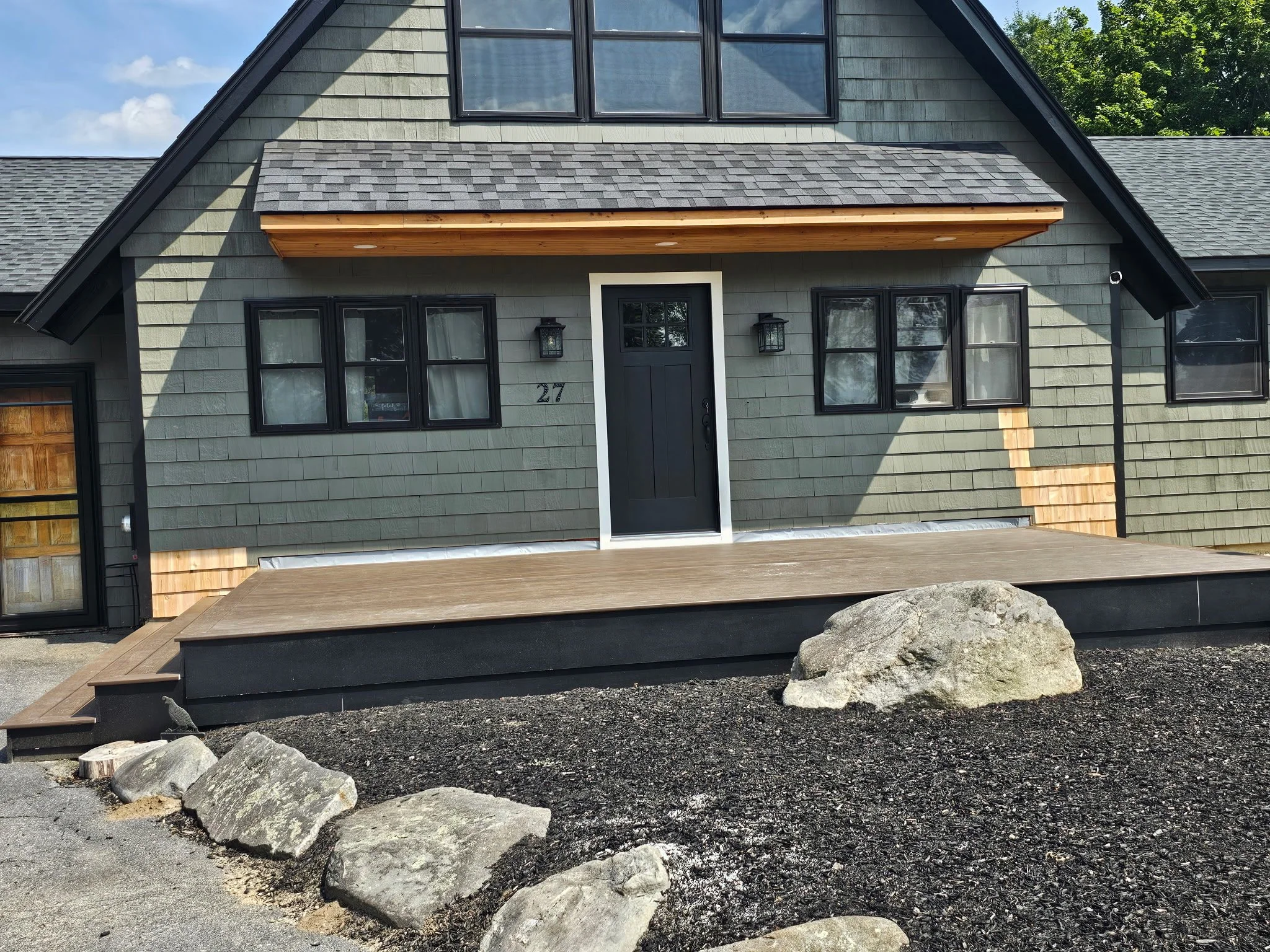Newly renovated house with gray siding, black window frames, a black front door, and a wooden porch. Rocks and dark mulch in the front yard.