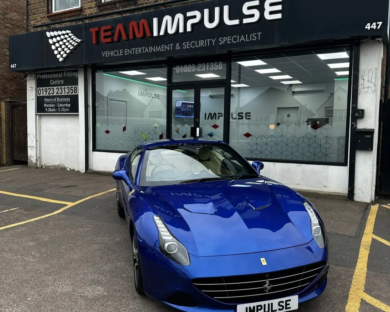 A blue Ferrari sports car parked outside a vehicle entertainment and security shop called Team Impulse. The shop has a large window with the company logo and signage, and the car is in a parking lot with yellow lines.