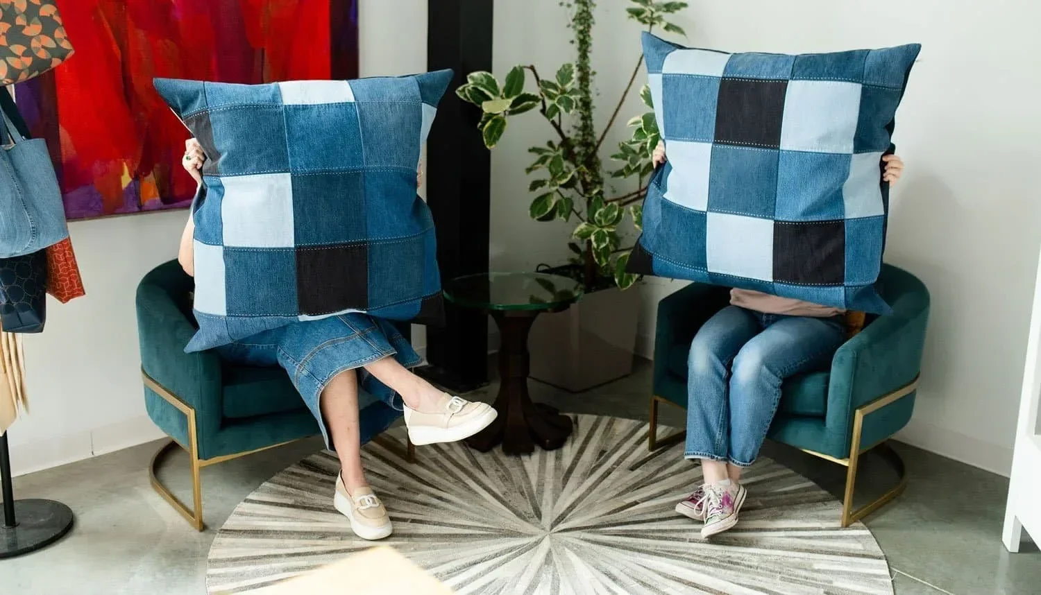 Two people sitting on teal chairs holding large blue checkered pillows in front of their faces, in a living room. One person is wearing white shoes and a denim skirt, the other is wearing sneakers and jeans.