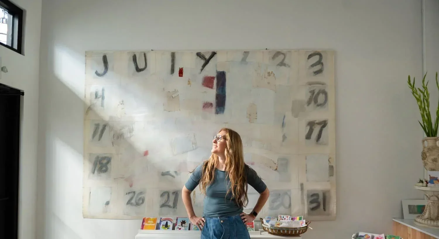 Tiffany (owner of Test Patch Studio) standing in front of large calendar artwork on a white wall, sunlight coming through a window on the left, with a plant on a white surface to the right.
