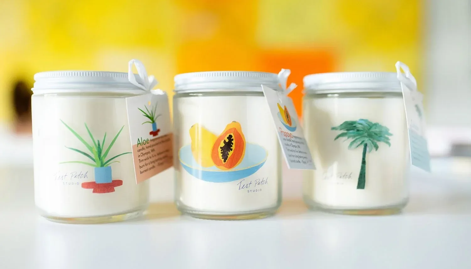 Three candles with illustrated labels featuring plants and fruits, including aloe, papaya, and a palm tree, placed on a white surface with a blurred colorful background.