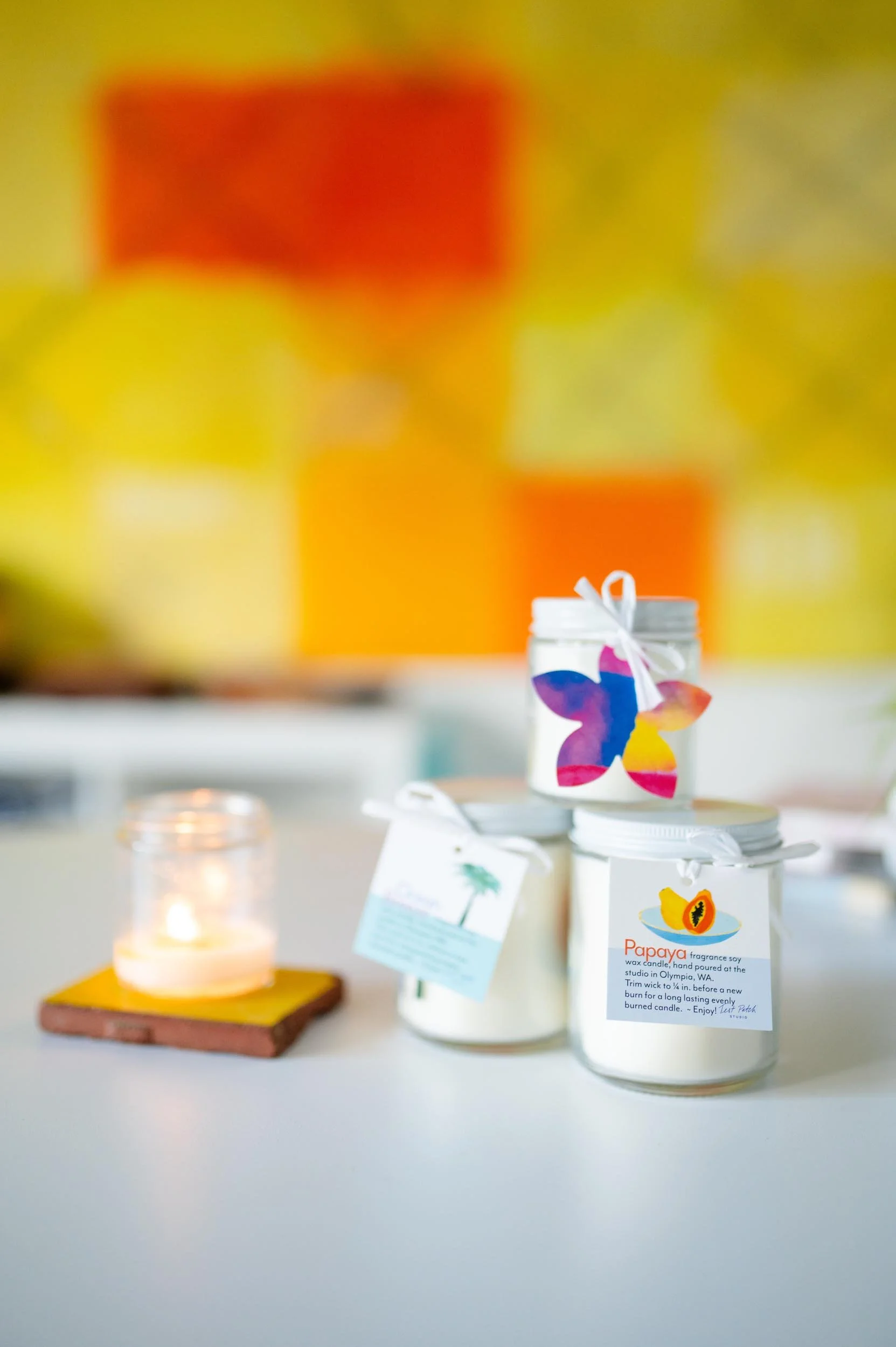 Jars of scented wax candles with butterfly and papaya labels on a white surface, with a lit candle in a glass container beside them and a colorful, abstract background.
