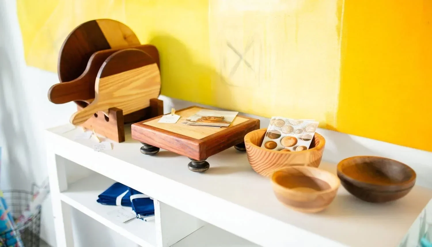 Display of handmade wooden bowls, cutting boards, and serving utensils on a white shelf against a yellow wall.