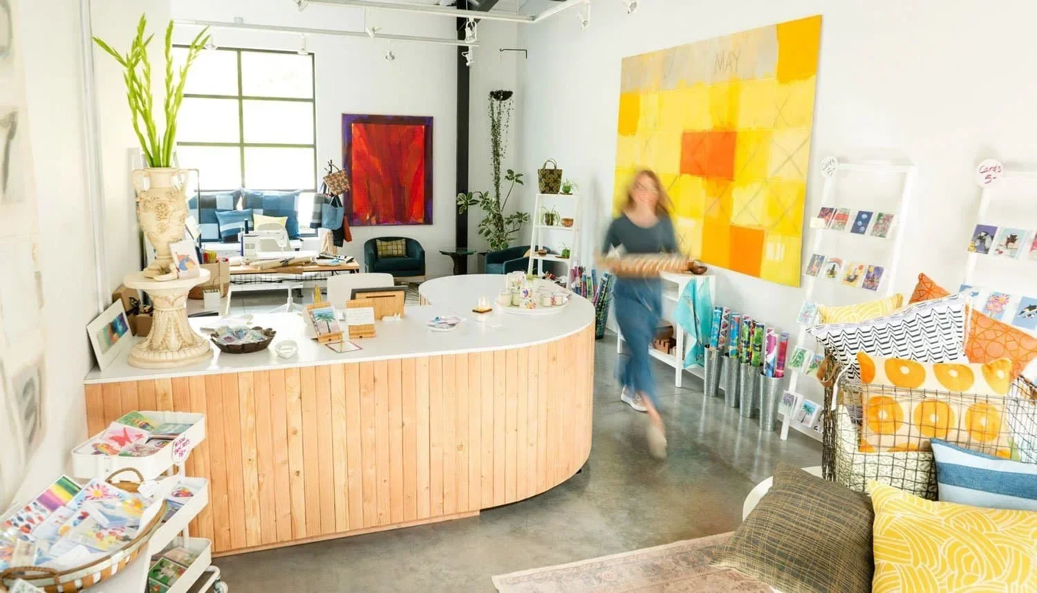 Interior of a brightly lit shop with a white curved counter made of wood in the foreground. Behind the counter, a blurred woman is holding a tray and walking. To the right, shelves display greeting cards and pillows with orange, yellow, and black-and