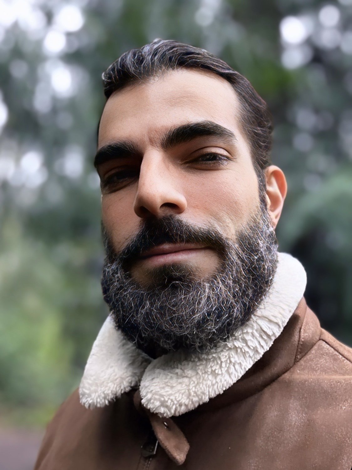 A close-up photo of a man with a beard and dark hair, wearing a brown jacket with a white, fluffy collar, outdoors with blurred trees in the background.
