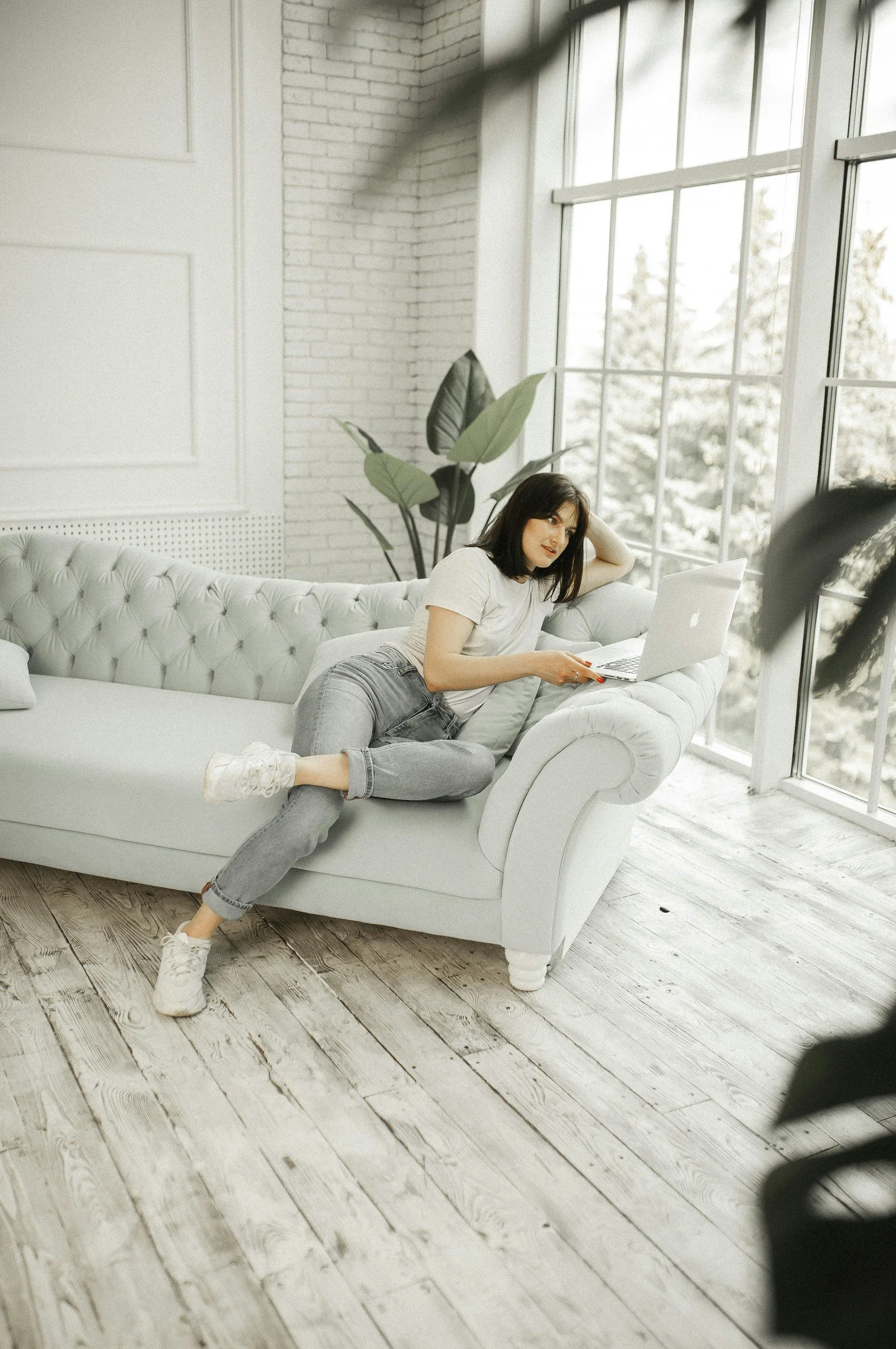 A woman relaxing on a white tufted sofa with a laptop in a bright room with large windows and potted plants.