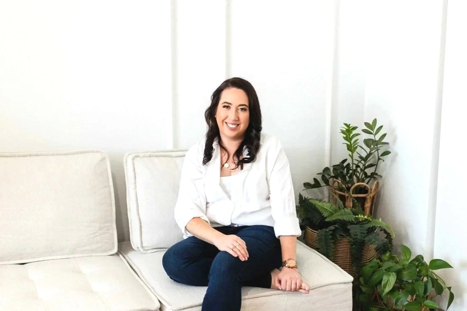 A woman with dark hair wearing a white blouse and blue jeans, sitting on a beige sofa with white cushions, smiling at the camera, with green potted plants behind her in a bright, minimalistic room.