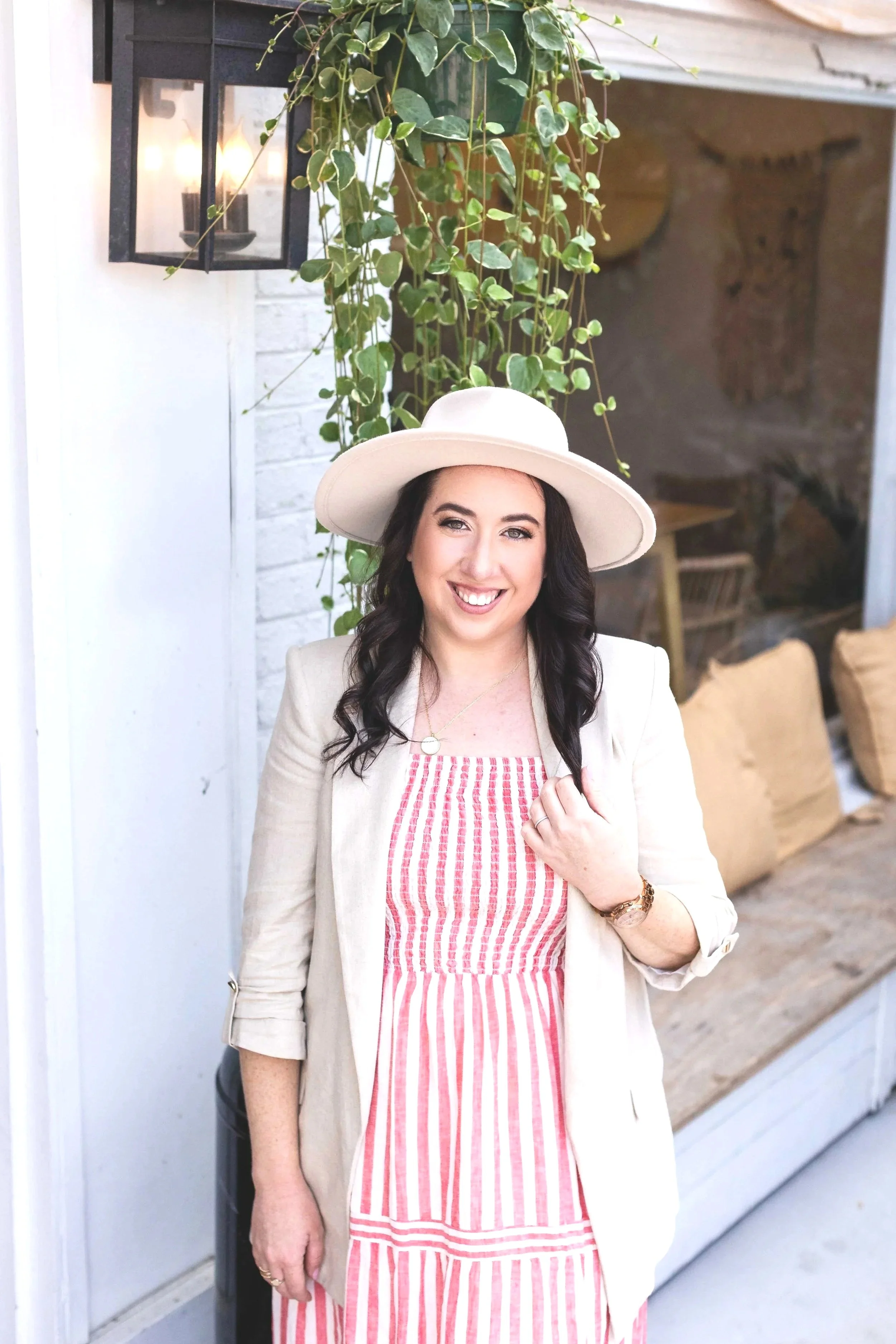 A woman with dark, wavy hair wearing a white wide-brimmed hat, a beige blazer, a pink and white striped dress, a gold wristwatch, necklace, and rings standing outdoors in front of a white brick wall, hanging greenery, and a lantern-style light fixture.