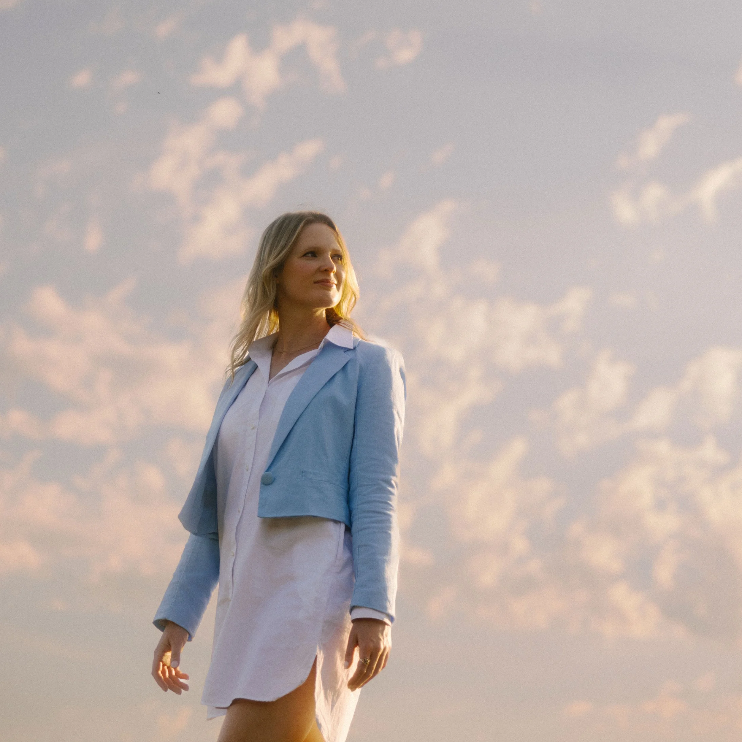 Somer Muhs, a licensed therapist, wearing a light blue blazer over a white dress walking outdoors during sunset with a cloudy sky in the background.